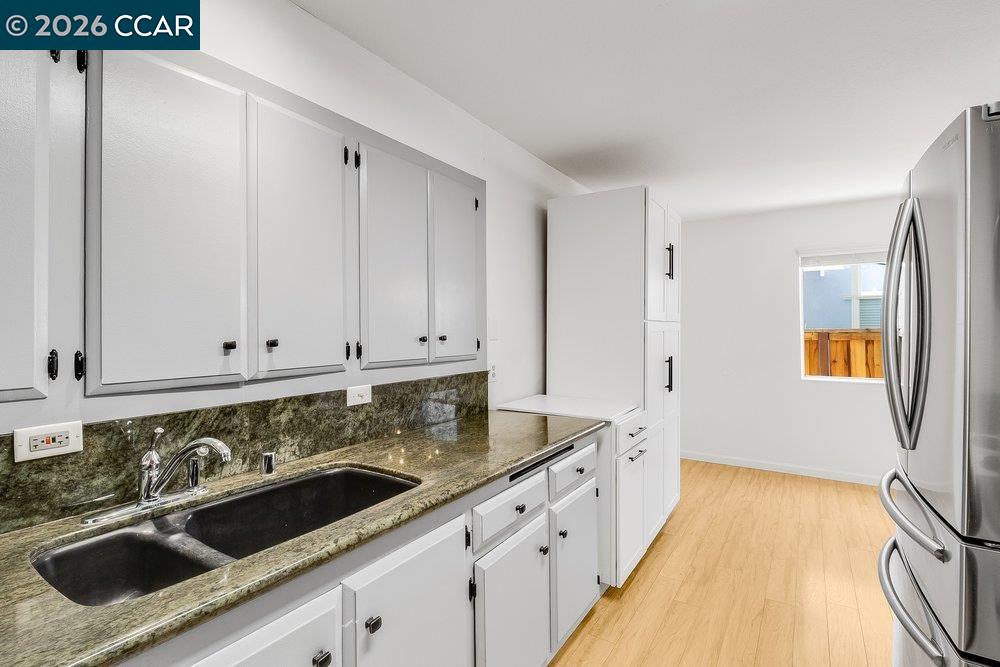 Kitchen with white cabinets, stone counters, double sink, stainless steel refrigerator, and light wood flooring.