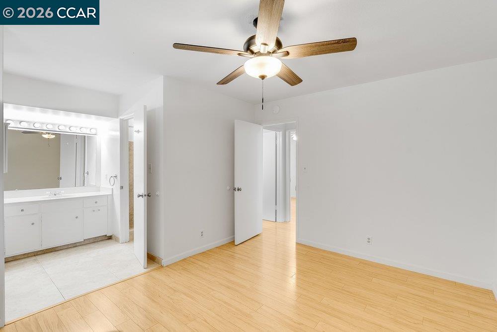 Empty bedroom with light wood floors and ceiling fan, adjoining a bathroom vanity.