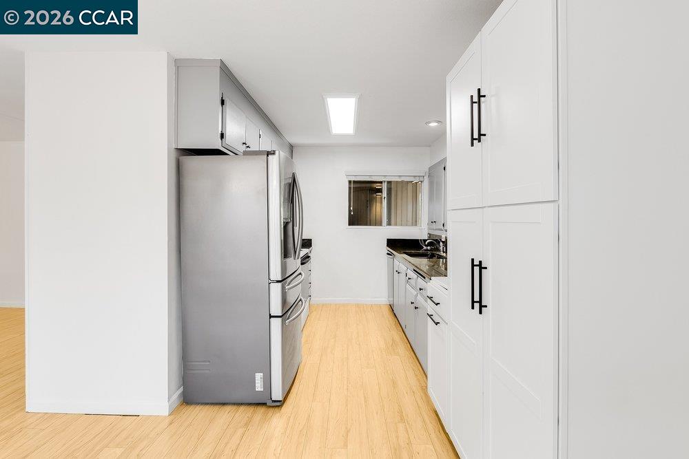 Galley kitchen with white cabinets, black counters, stainless steel refrigerator, and light wood flooring.