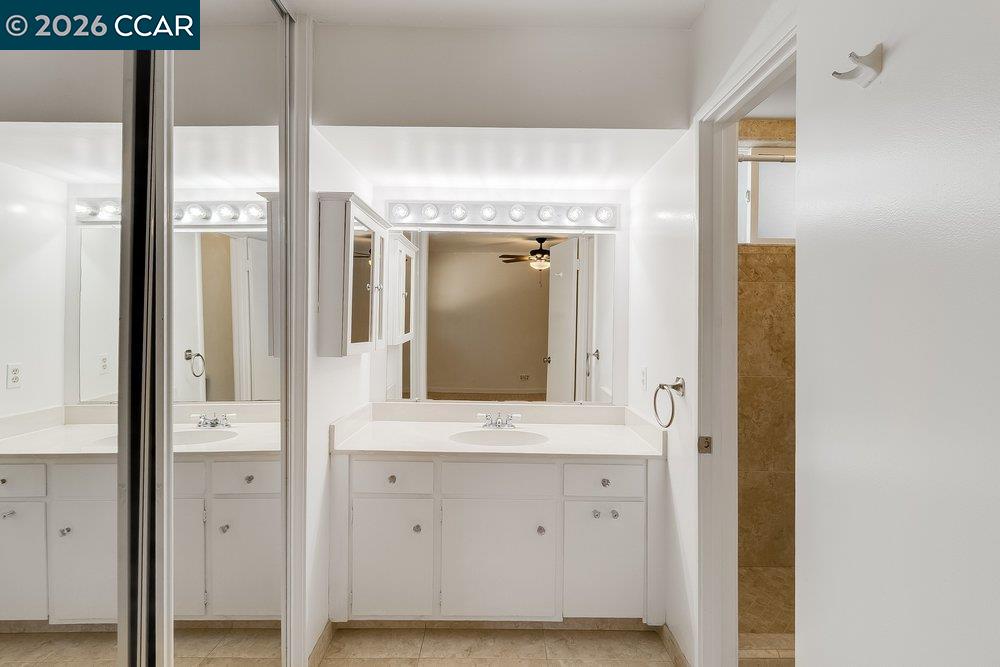Bathroom vanity with a wide mirror, white cabinets, bright lights, and a tiled shower doorway.