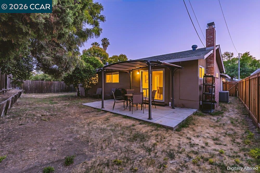 Rear exterior with covered patio, outdoor dining set, and fenced yard at dusk.