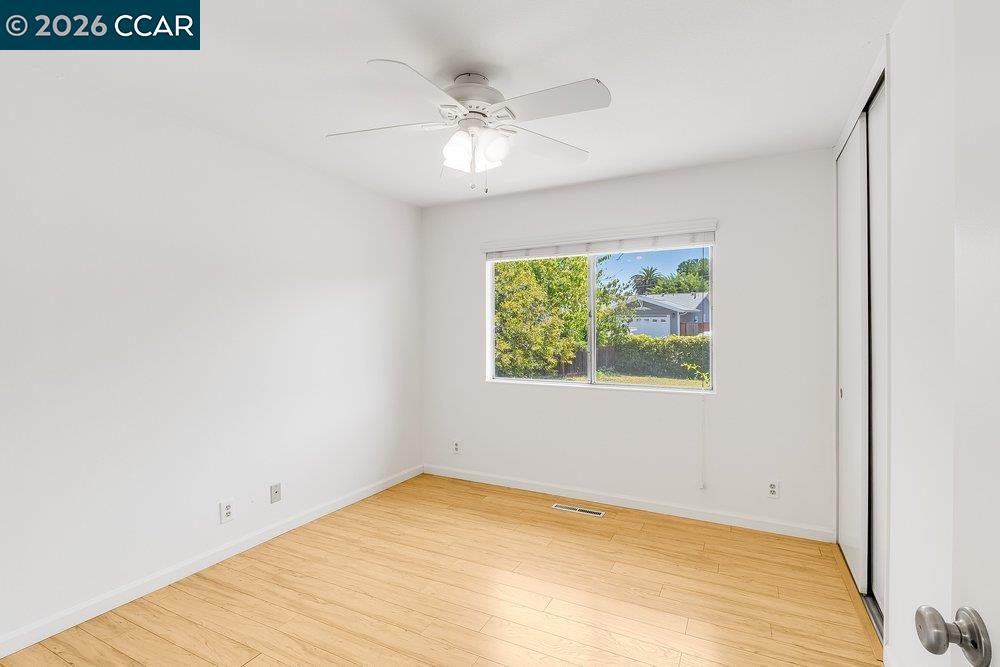 Empty bedroom with light wood flooring, a ceiling fan, and a window overlooking greenery.