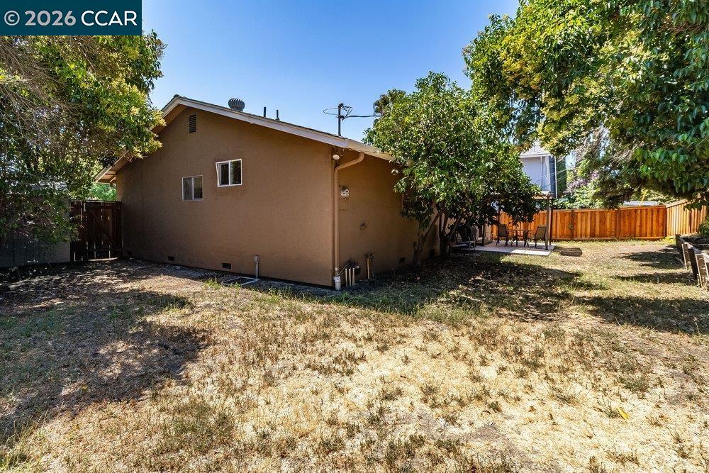 Tan house viewed from a dry backyard, with mature trees, a wooden fence, and patio seating.