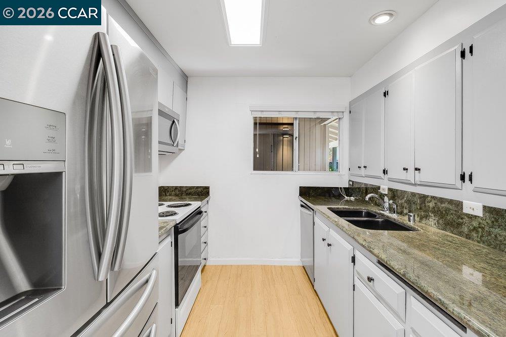 Galley kitchen with white cabinets, stainless steel appliances, dark patterned counters, and light wood flooring.