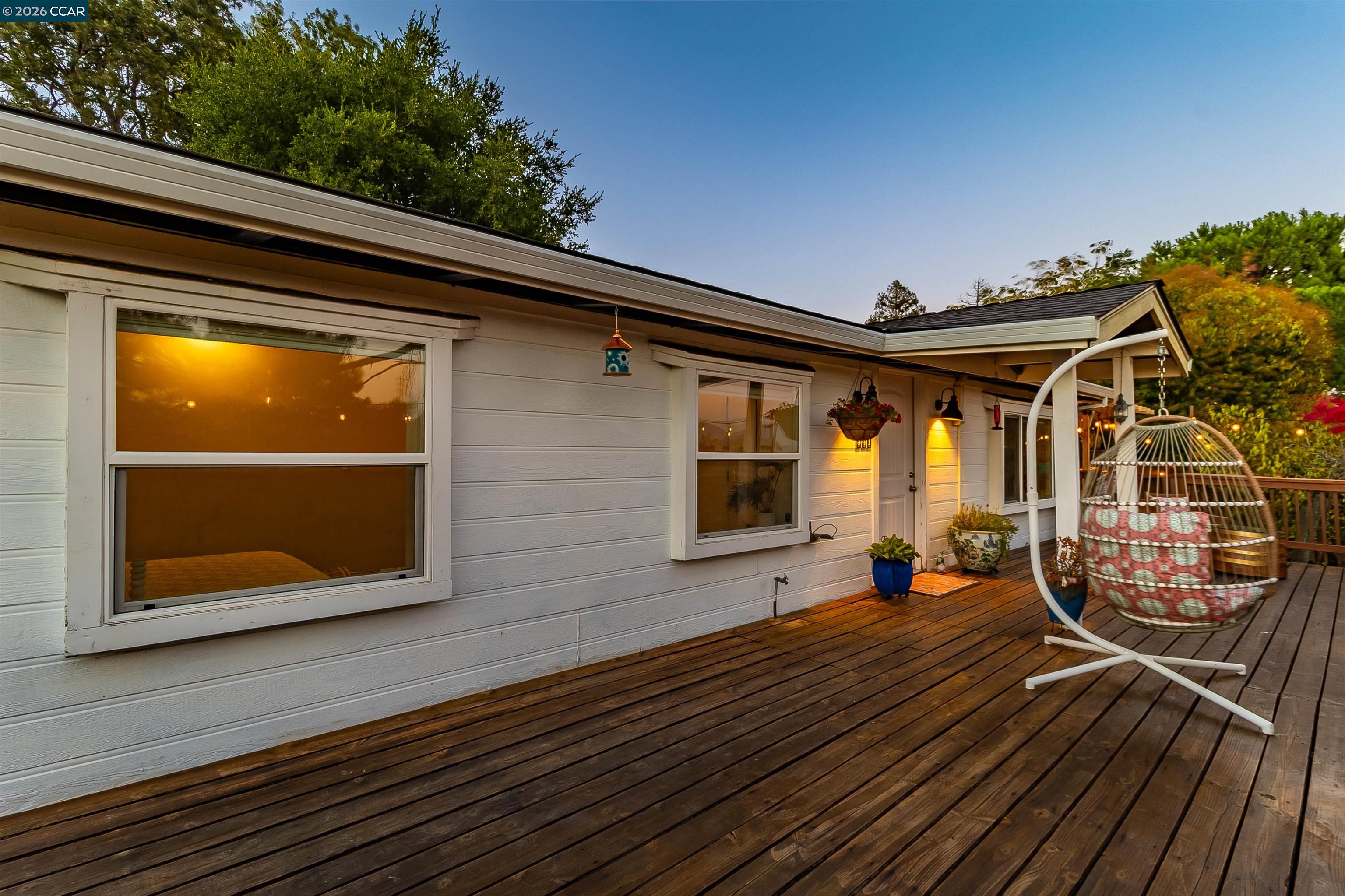 House exterior and wide wooden deck with a hanging chair and warm lights at dusk.