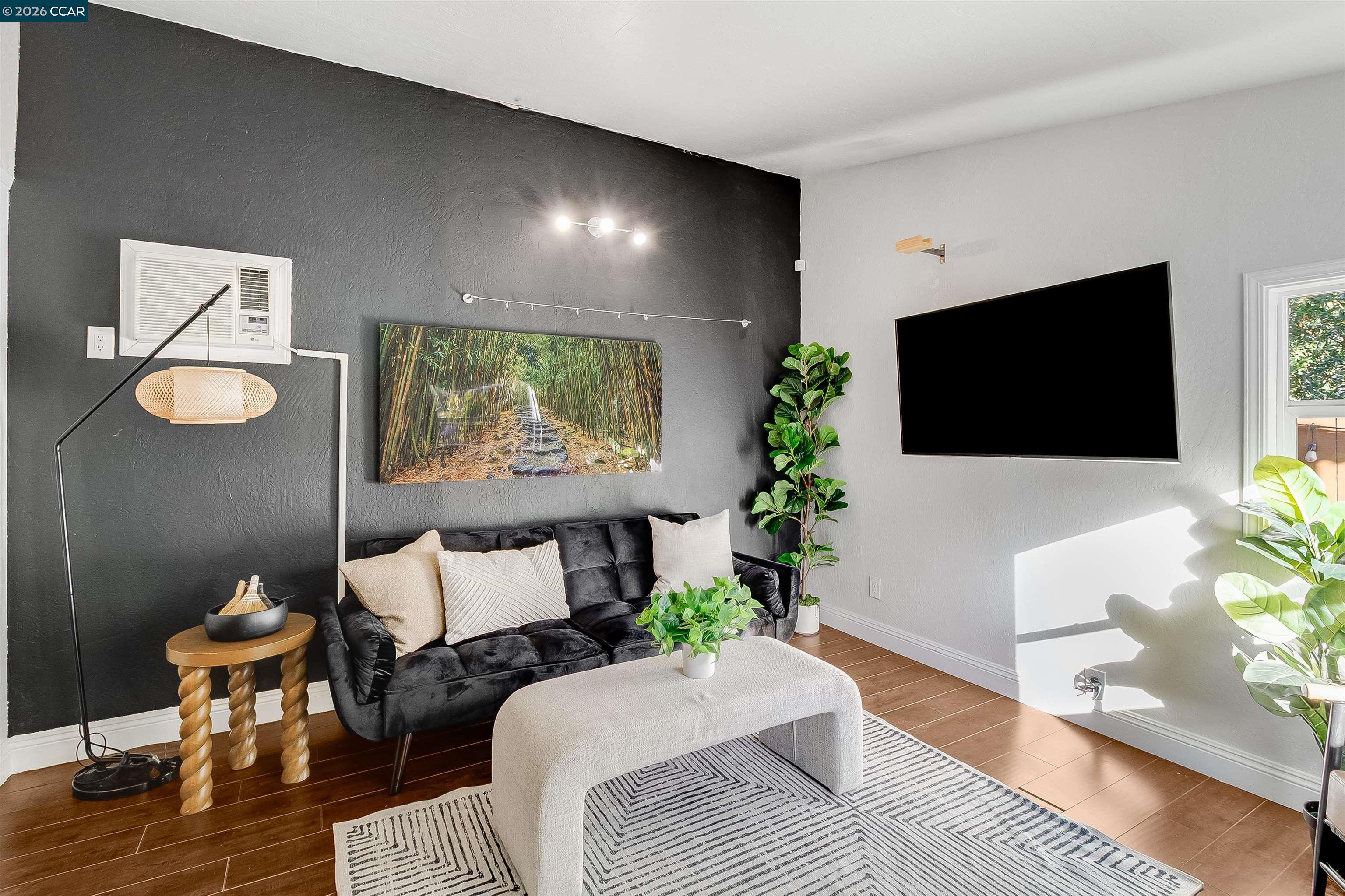 Furnished living room with a charcoal accent wall, black sofa, light ottoman, and wall-mounted television.