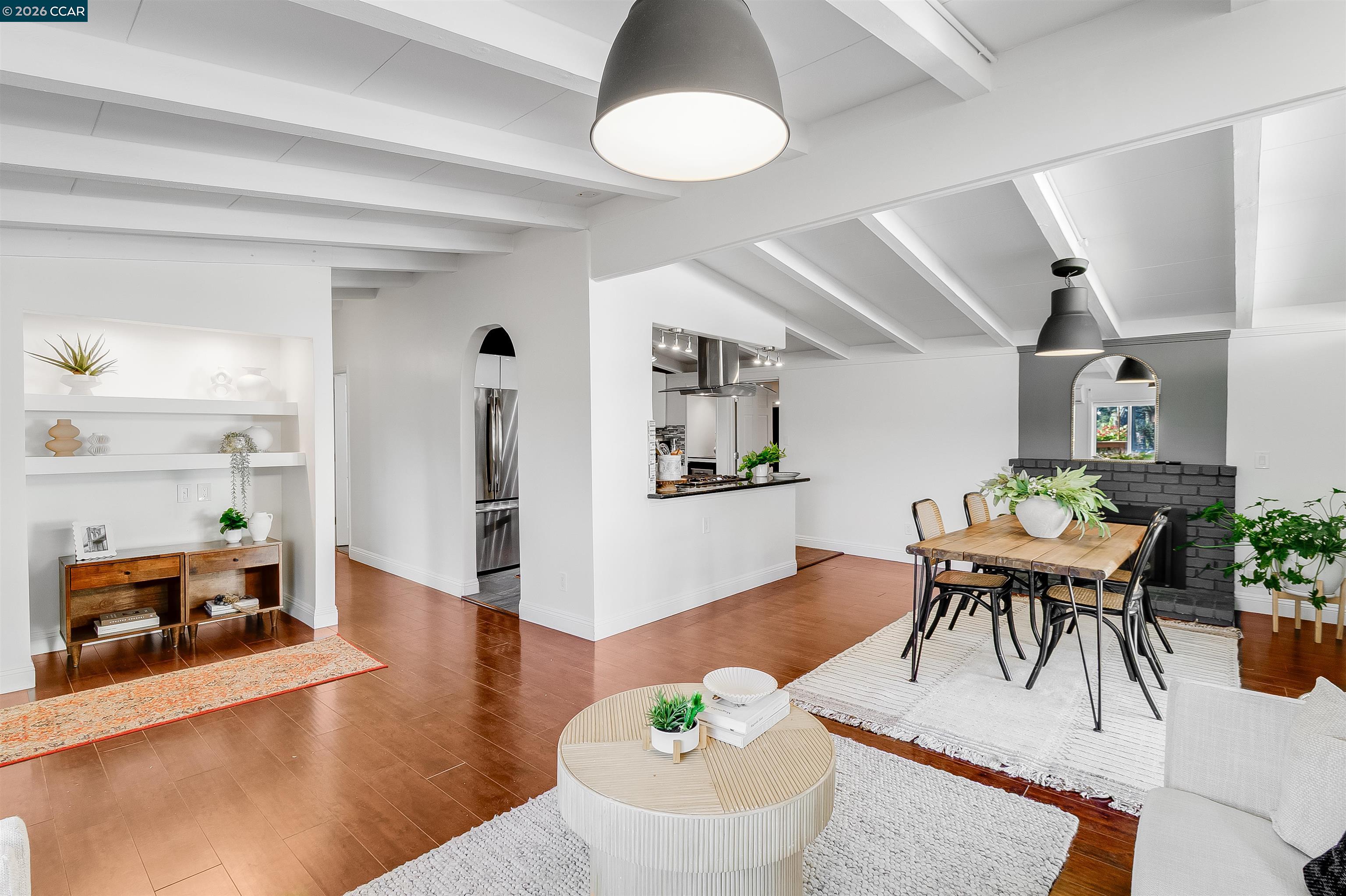 Open-plan living and dining room with hardwood floors, white ceiling beams, and a black brick fireplace.