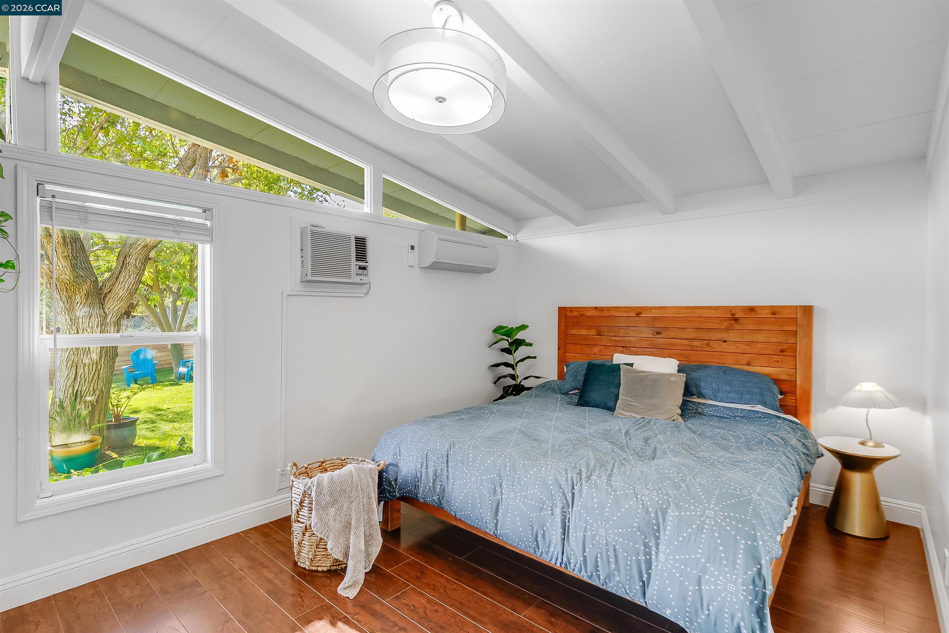 Bedroom with a wood-plank headboard, vaulted beamed ceiling, and garden view through a large window.