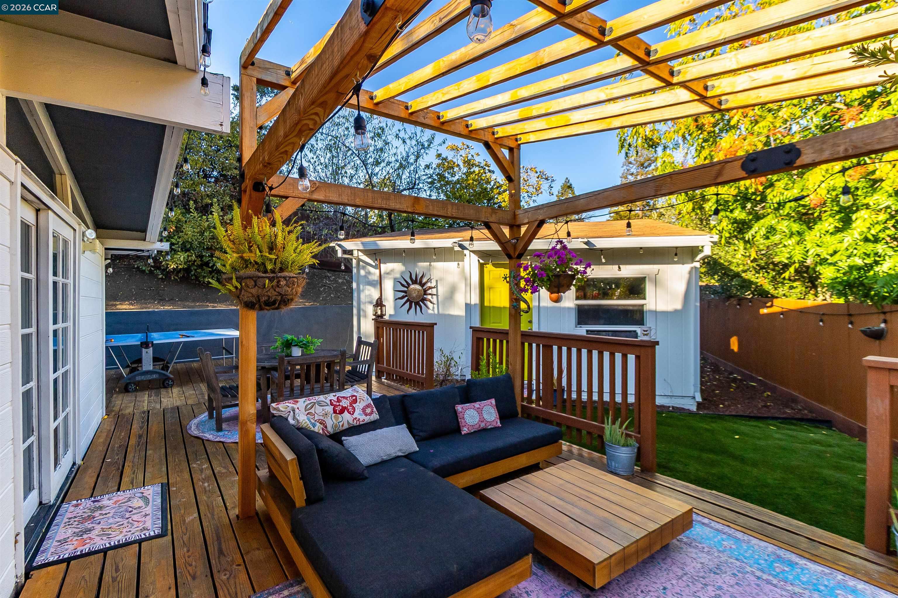 Backyard deck beneath a wooden pergola, with sectional seating, string lights, and a white shed with a yellow door.