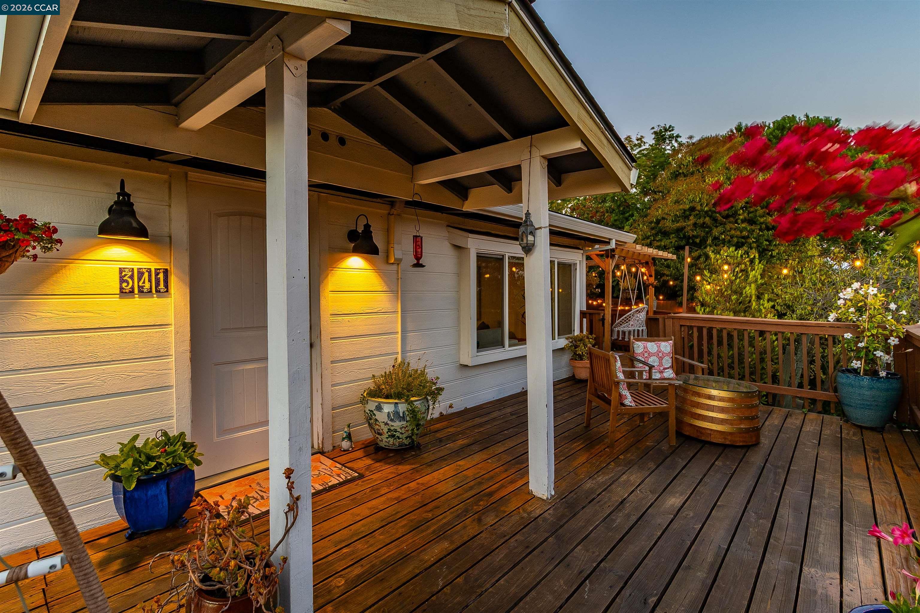 Covered wooden porch and deck with patio seating, potted plants, and warm lights at dusk.