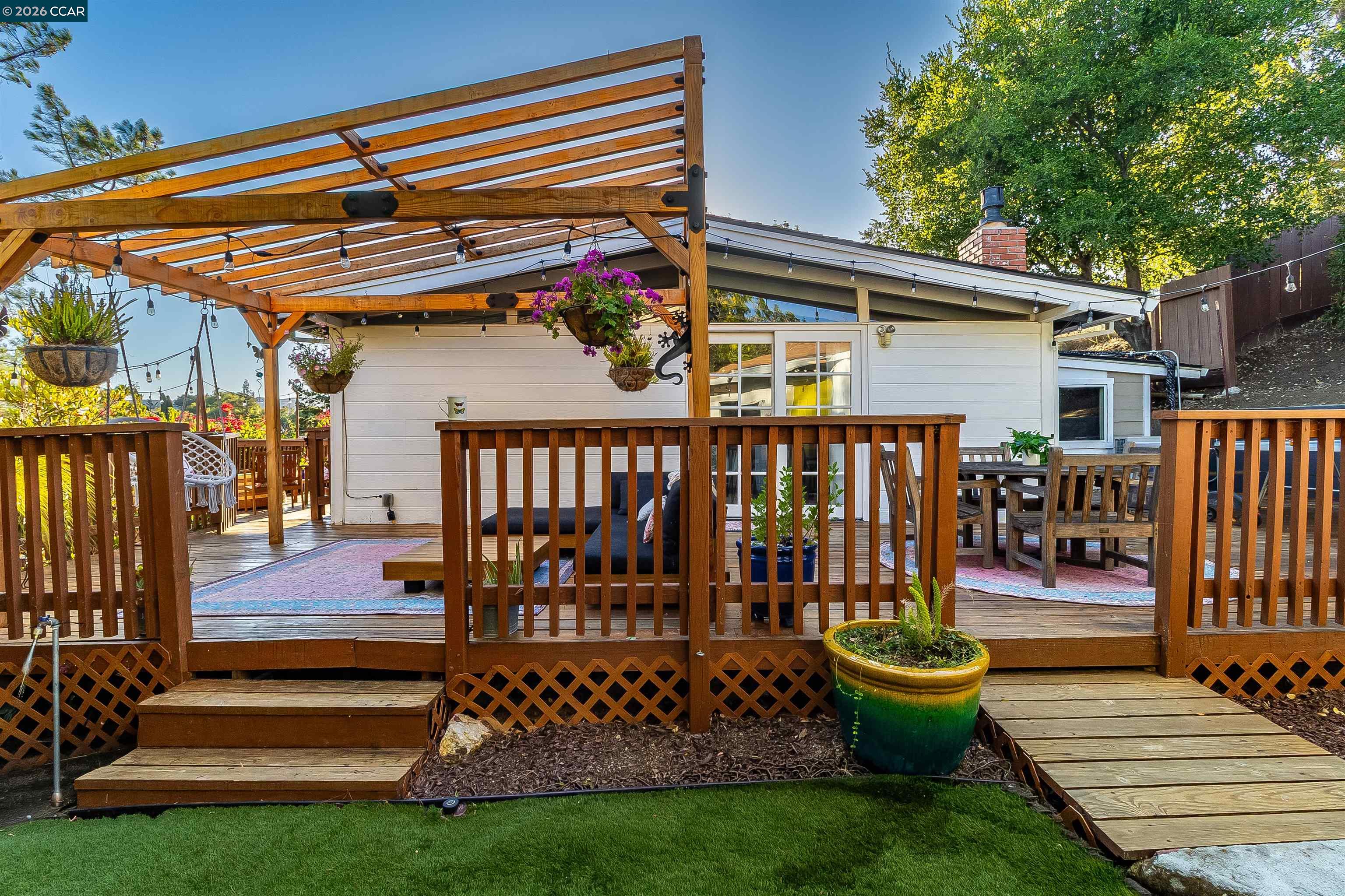 Rear deck with a wooden pergola, string lights, outdoor seating, and hanging flowers.