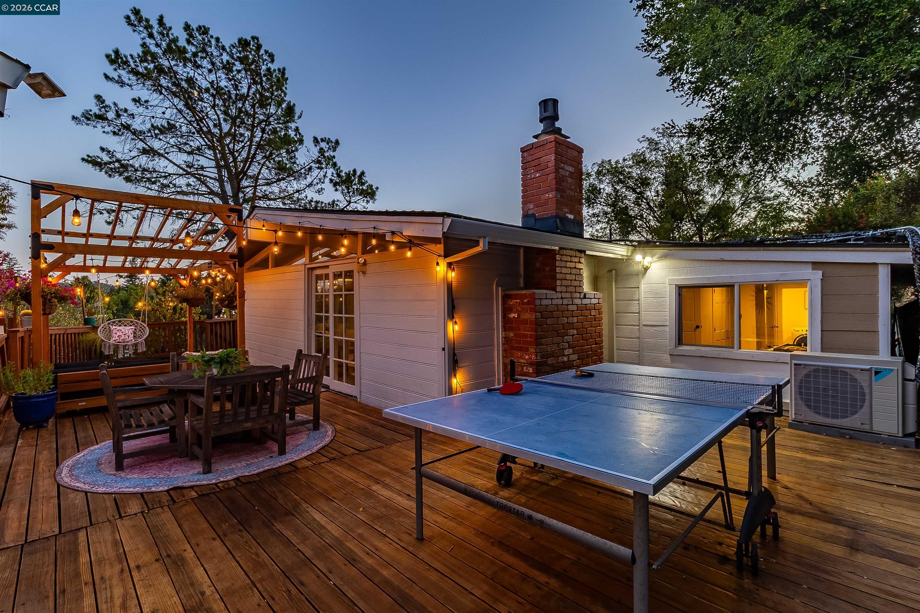 Backyard deck with a lit pergola, outdoor dining set, and ping-pong table at dusk.
