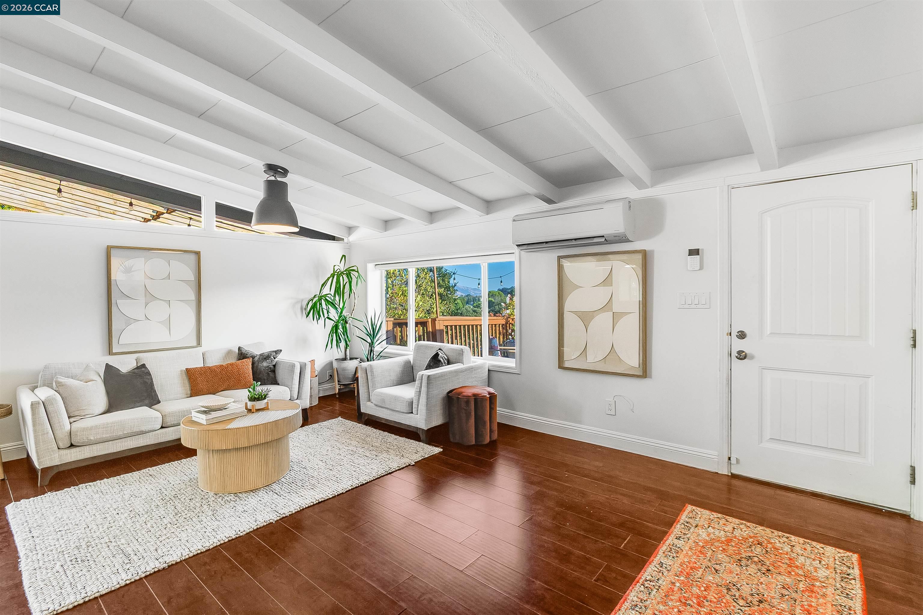 Furnished living room with white exposed beams, dark wood floors, and a window overlooking a deck.