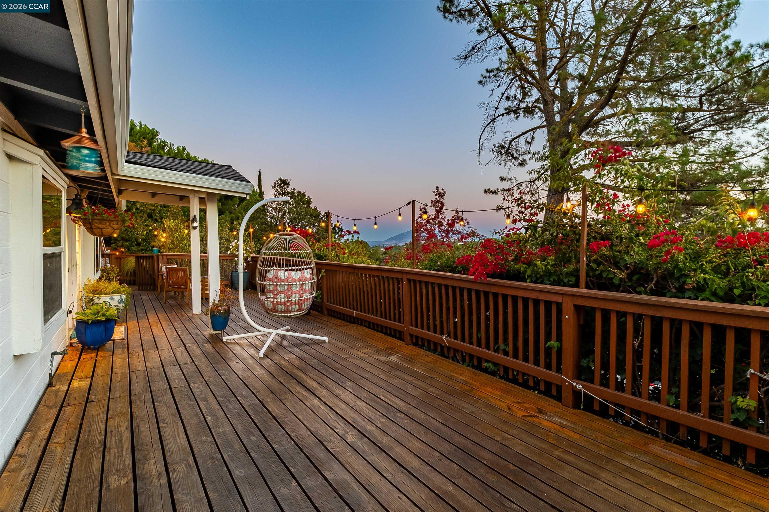 Wooden deck with a hanging egg chair, flowering shrubs, string lights, and distant hills at dusk.