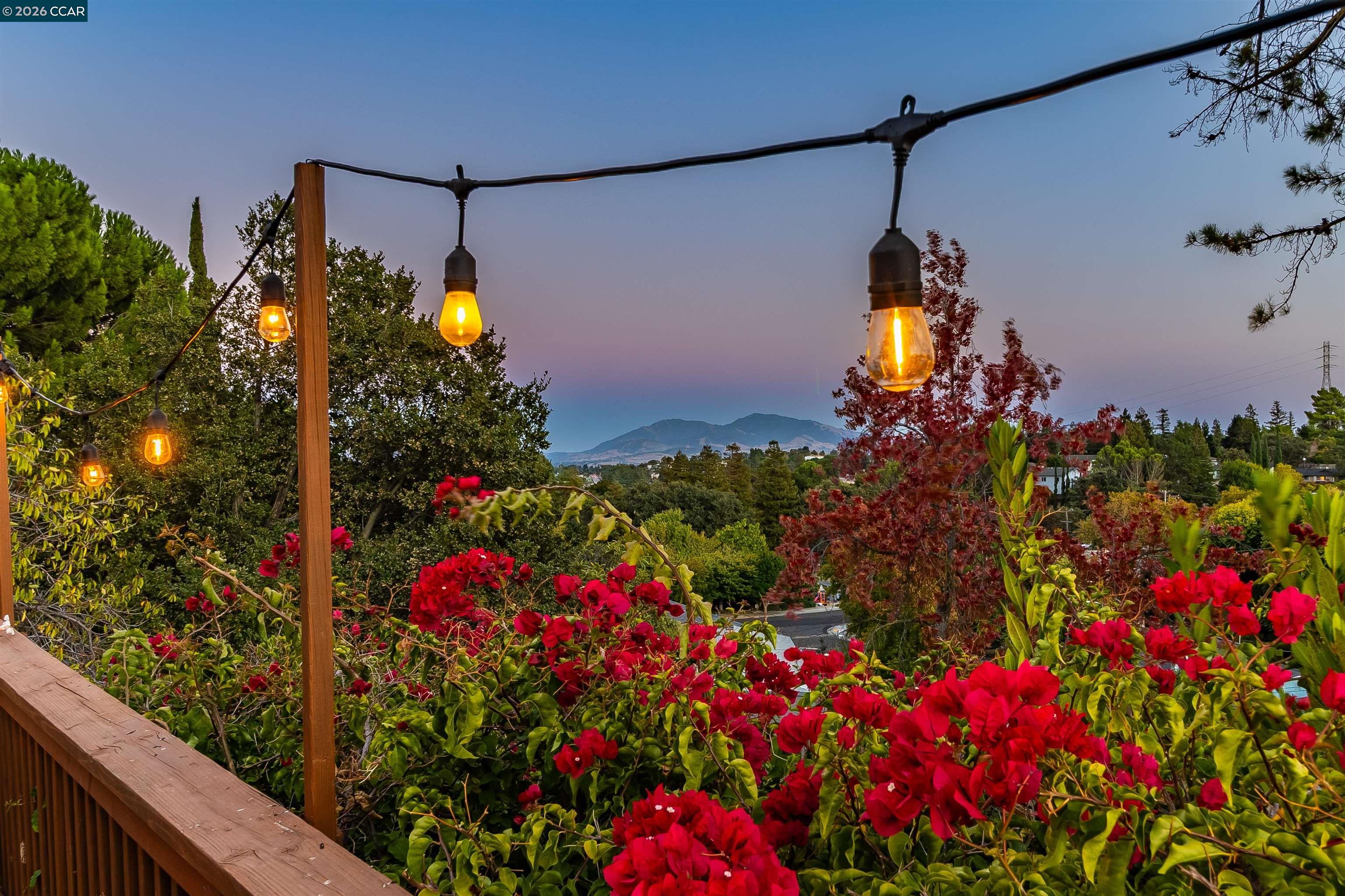 Deck overlooking distant hills at dusk, framed by glowing string lights and red bougainvillea.