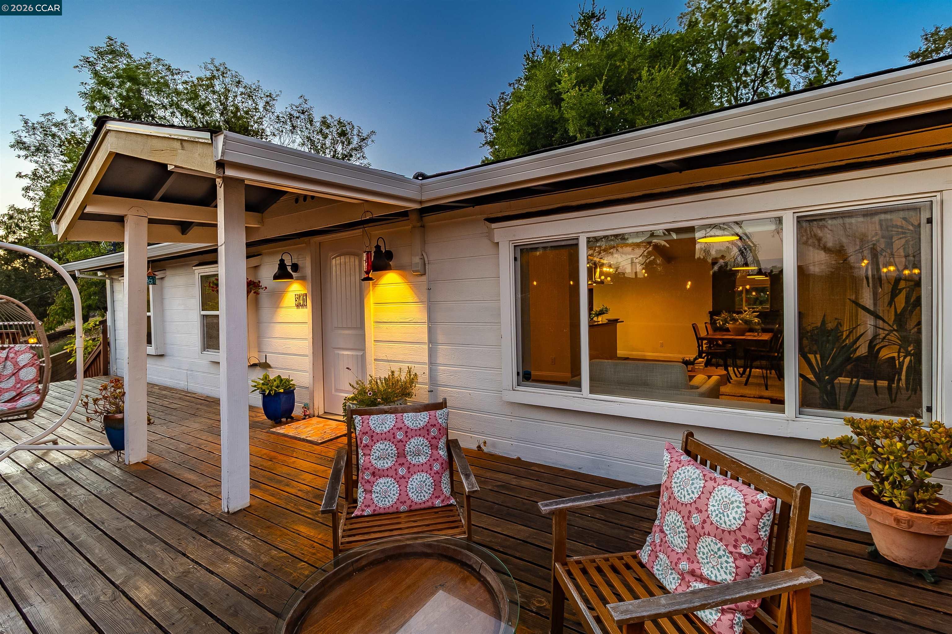 White house with a covered wooden deck, cushioned chairs, and warm lights glowing at dusk.