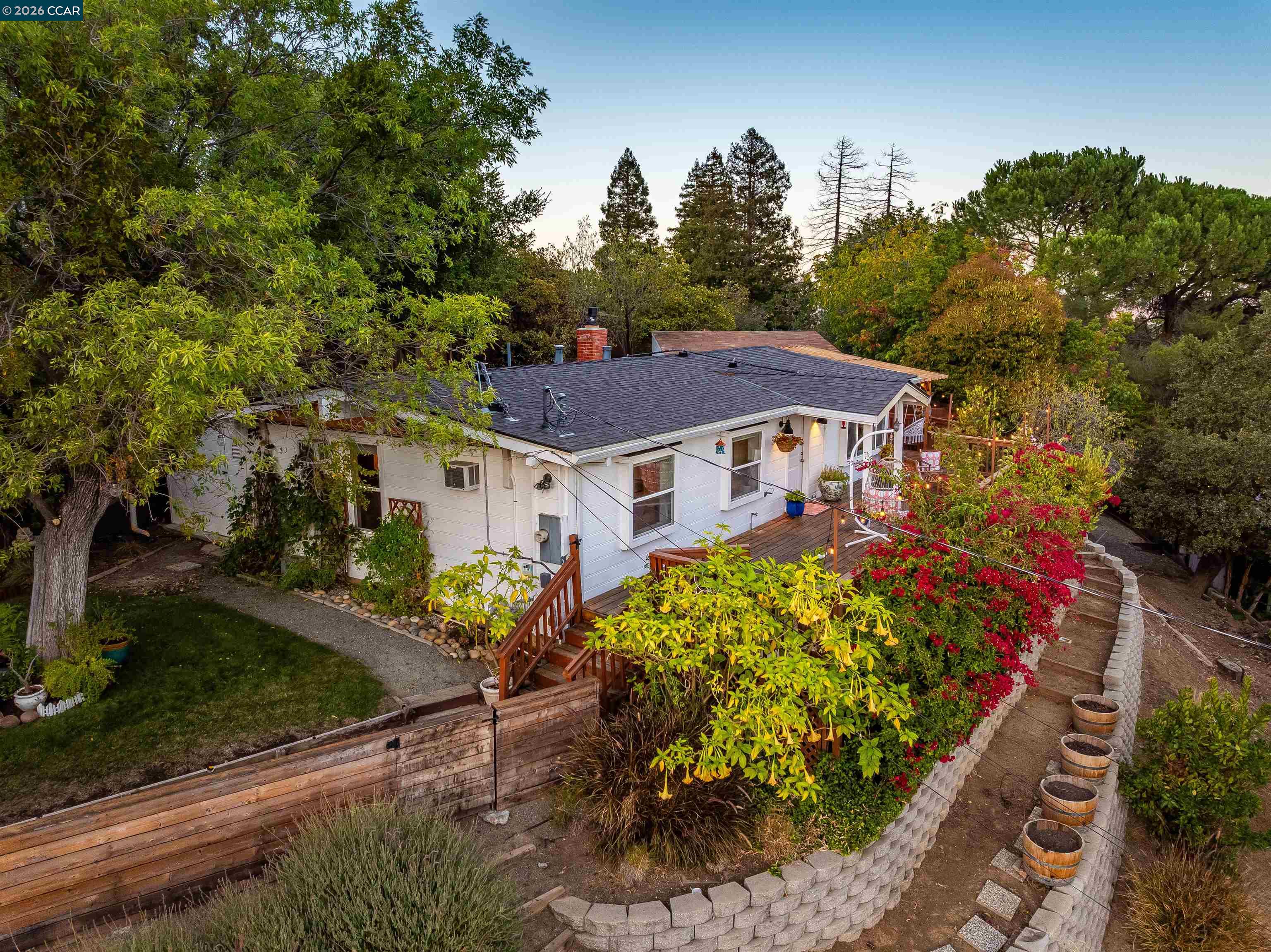 Aerial view of a white single-story home with a wooden deck, garden, and curved retaining wall.