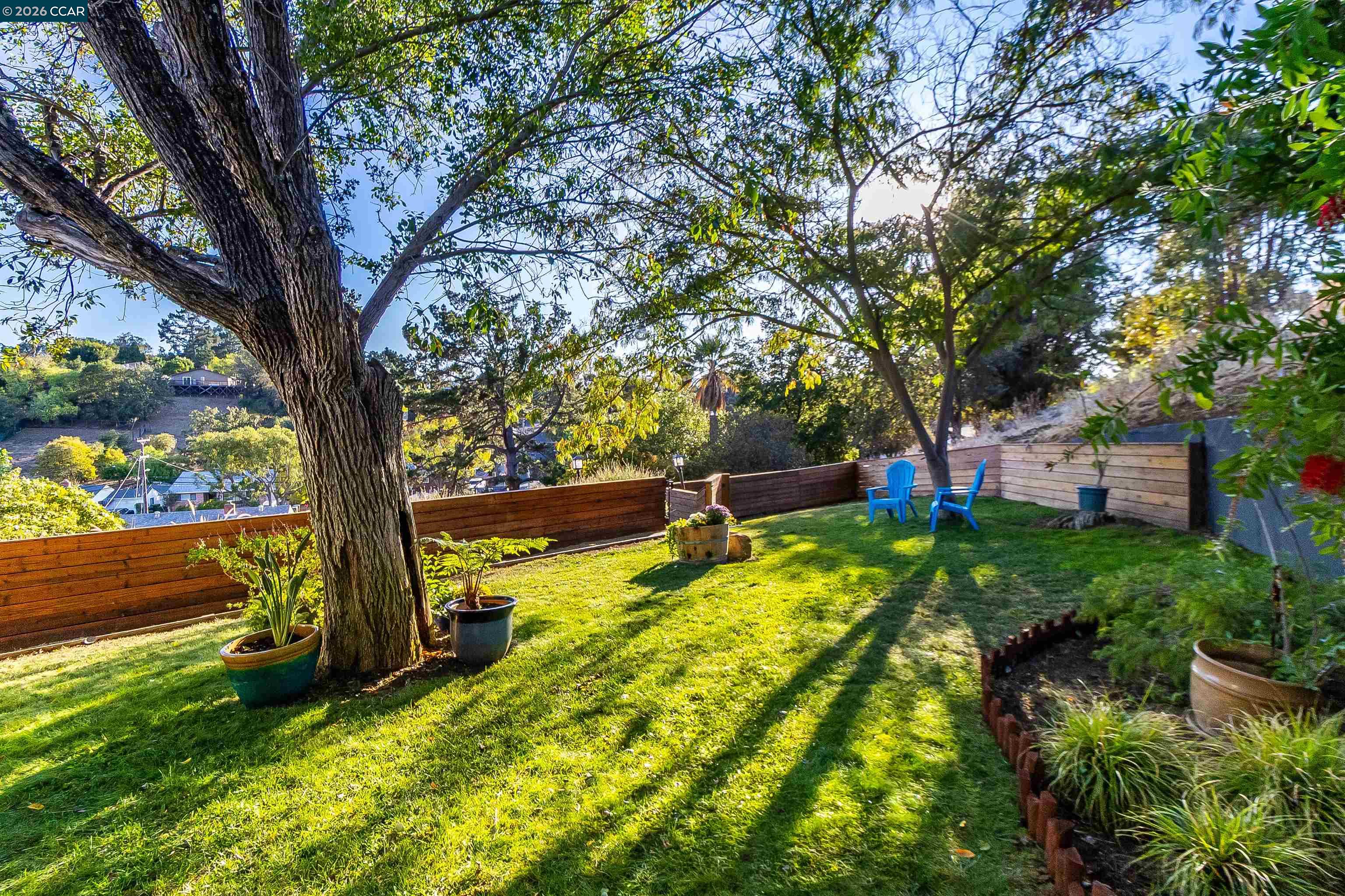 Fenced backyard with a green lawn, mature shade trees, and two blue Adirondack chairs.