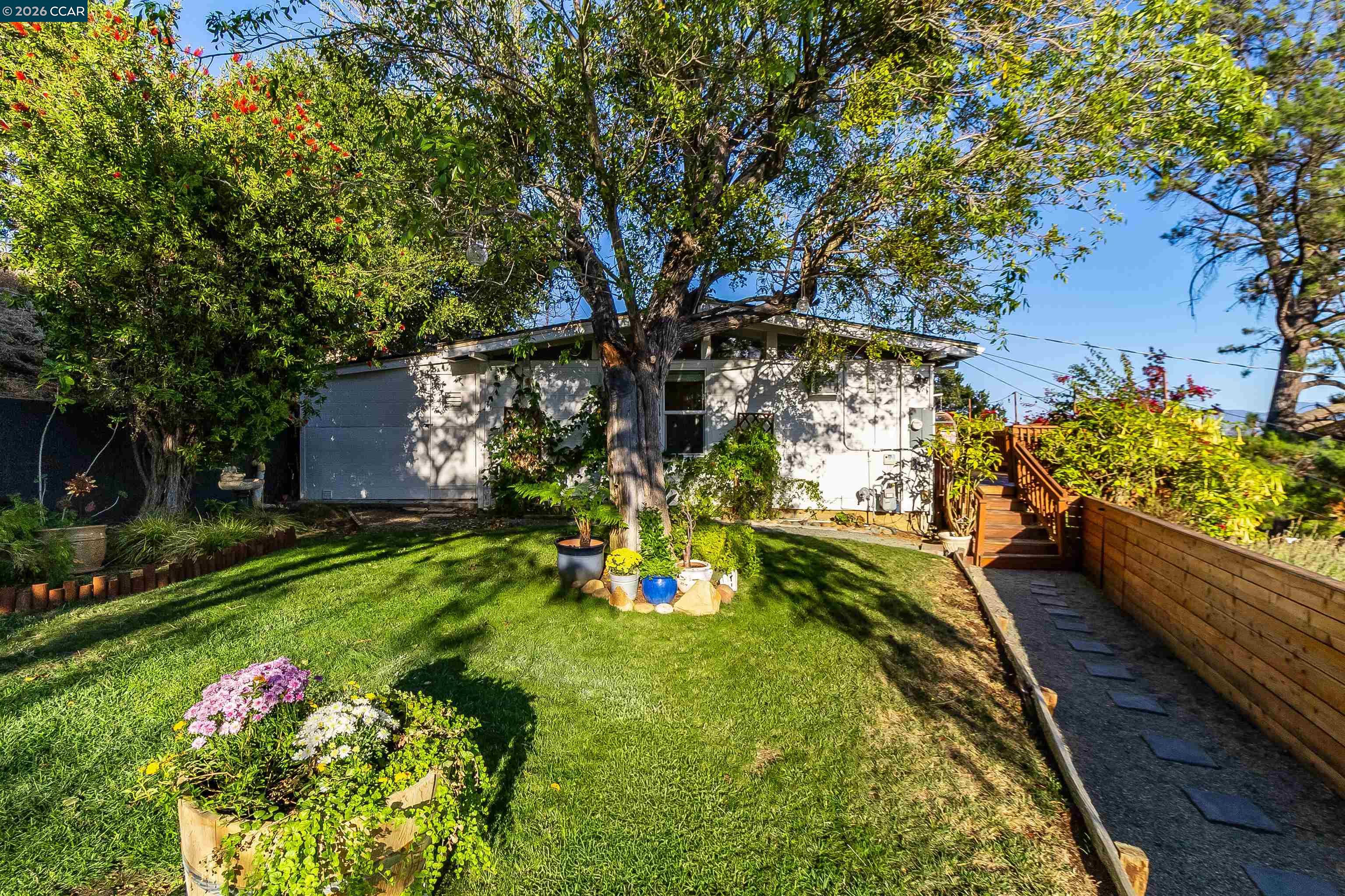 Rear of a single-story home beneath mature trees, with a lawn, flower beds, and wooden deck steps.