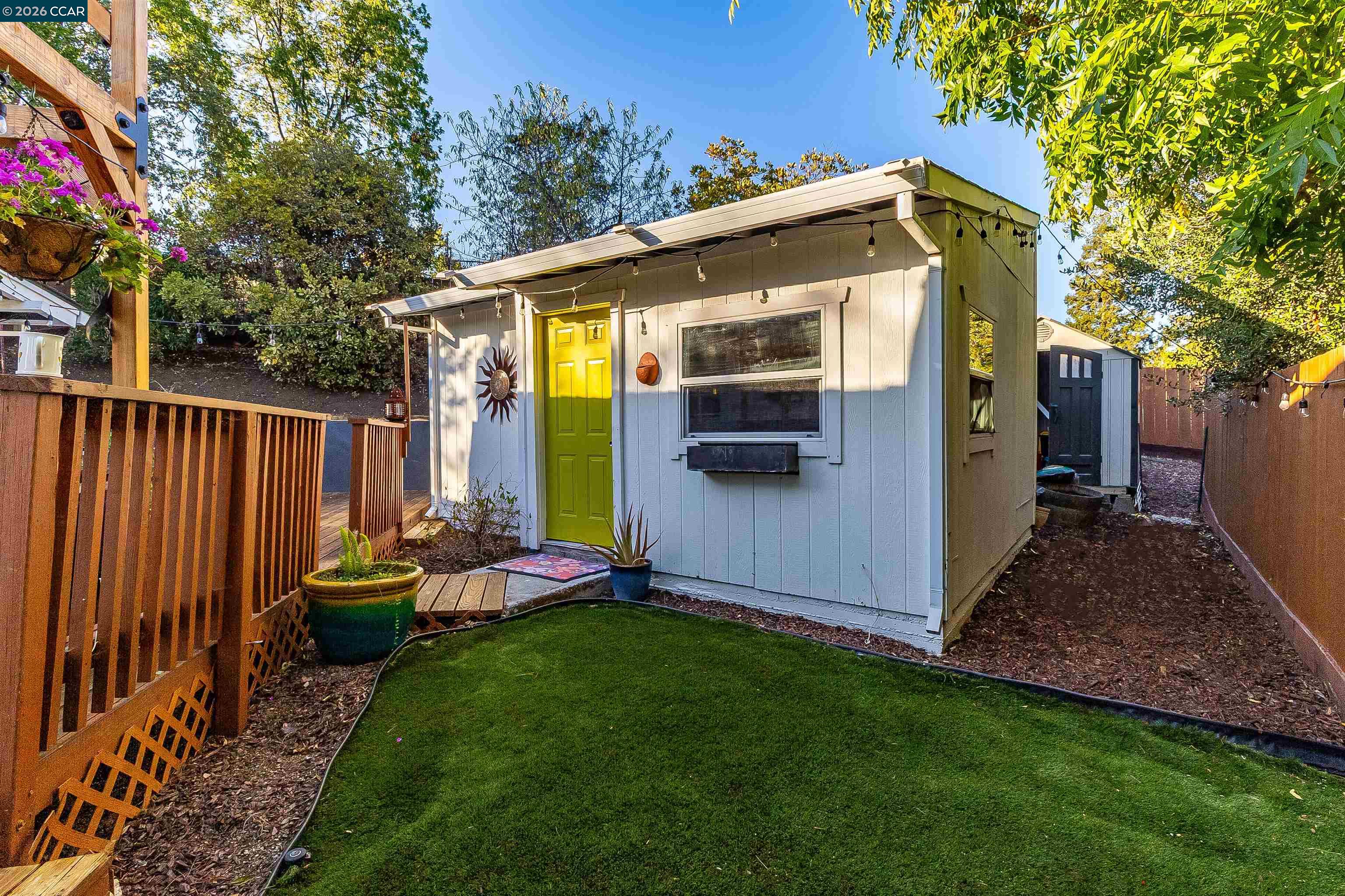 Backyard outbuilding with a bright yellow-green door, string lights, wooden deck, and artificial turf.
