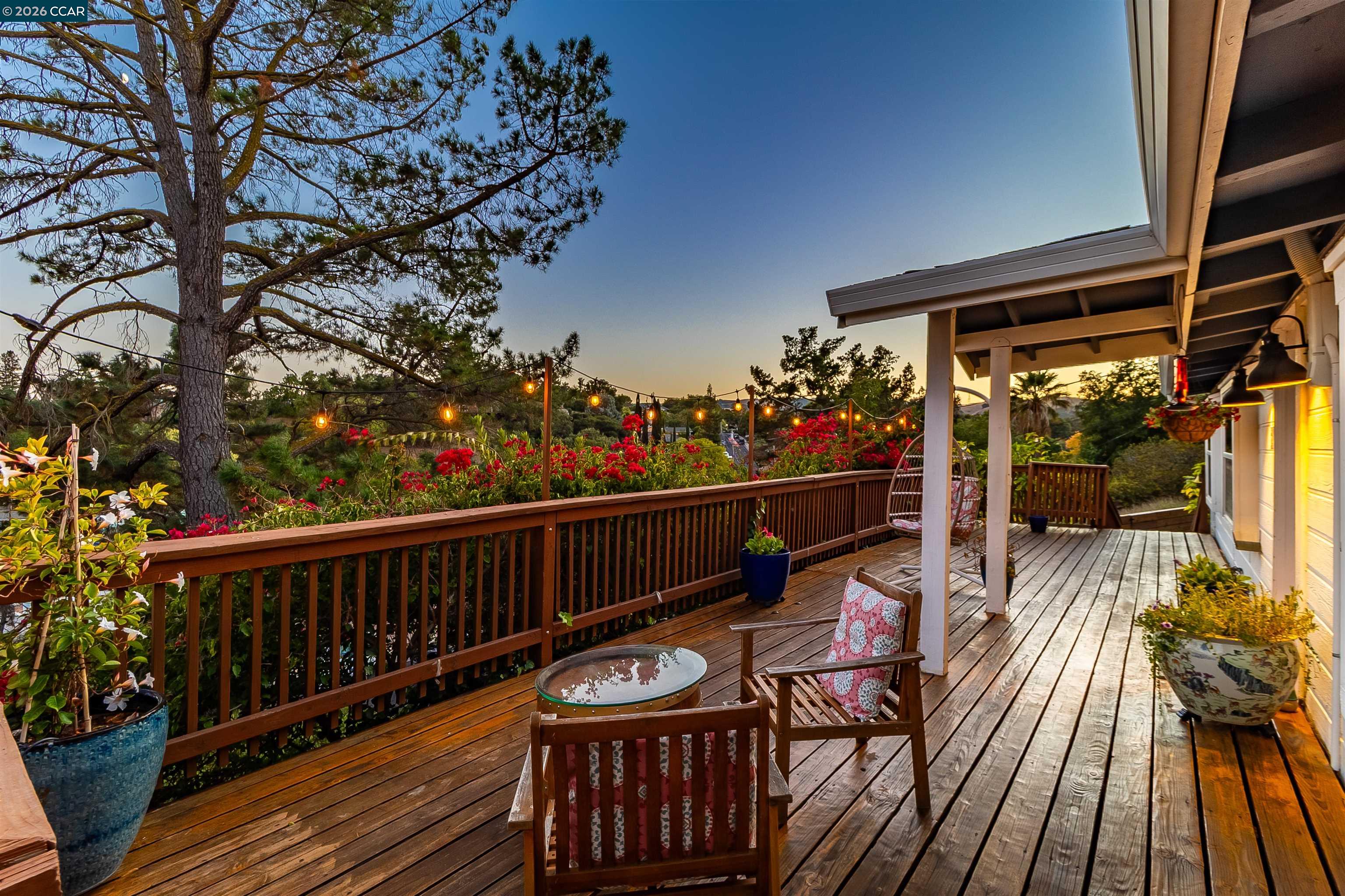 Wooden deck with patio seating, flowering shrubs, and string lights at dusk.