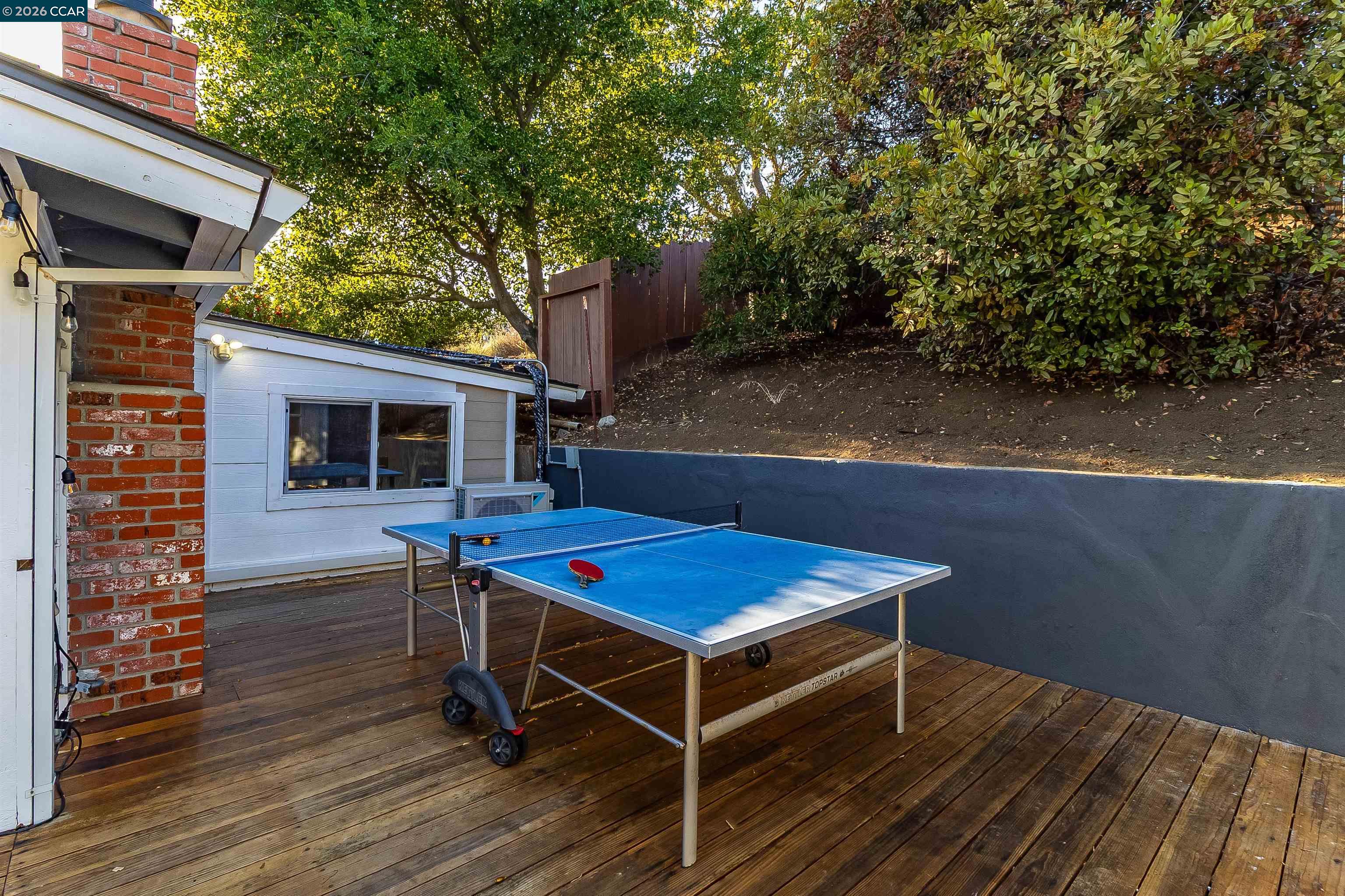 Backyard deck with a blue ping-pong table beside the house and a leafy hillside.