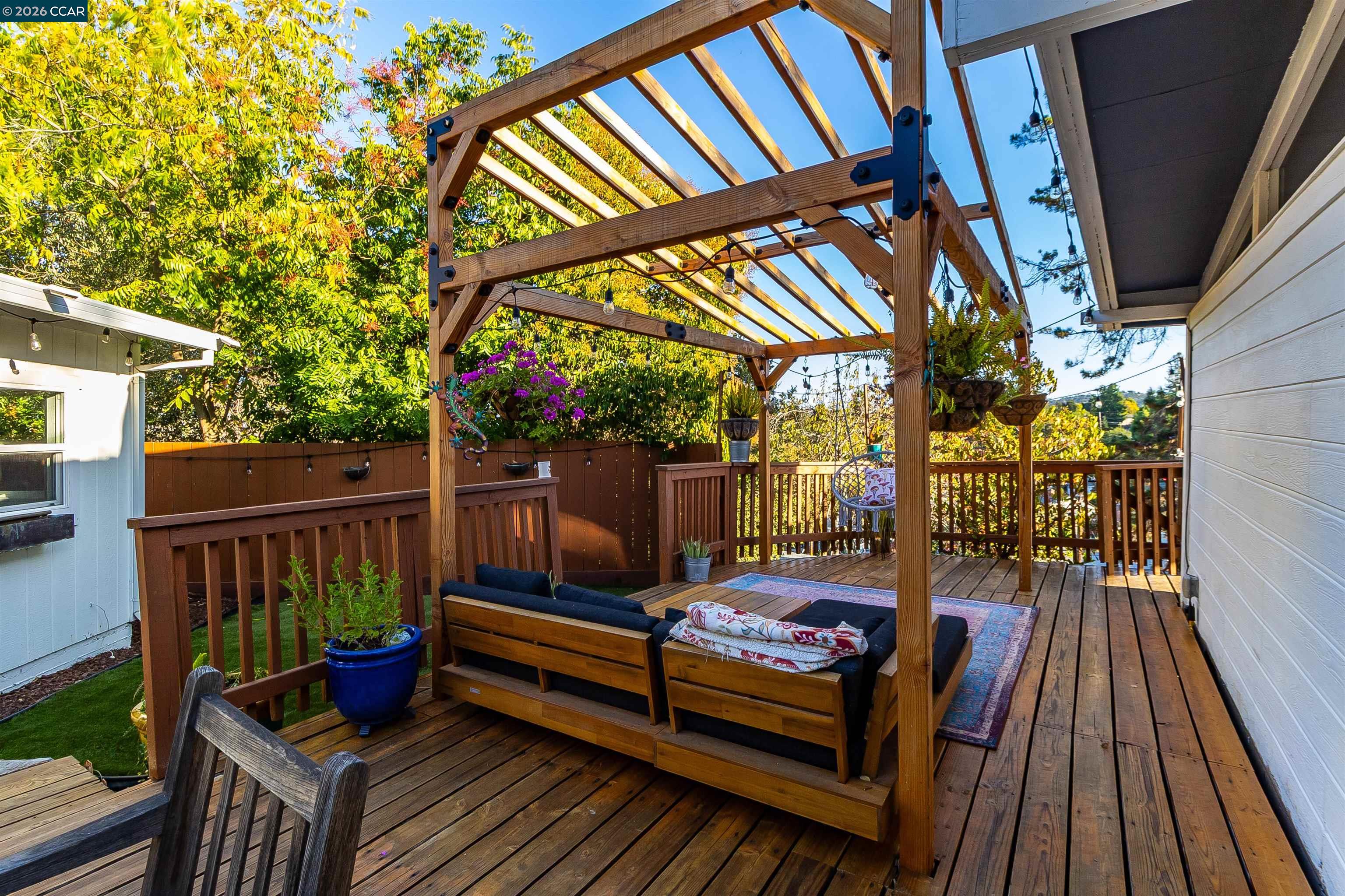 Wooden deck with a timber pergola, outdoor sofa, potted plants, and leafy trees.