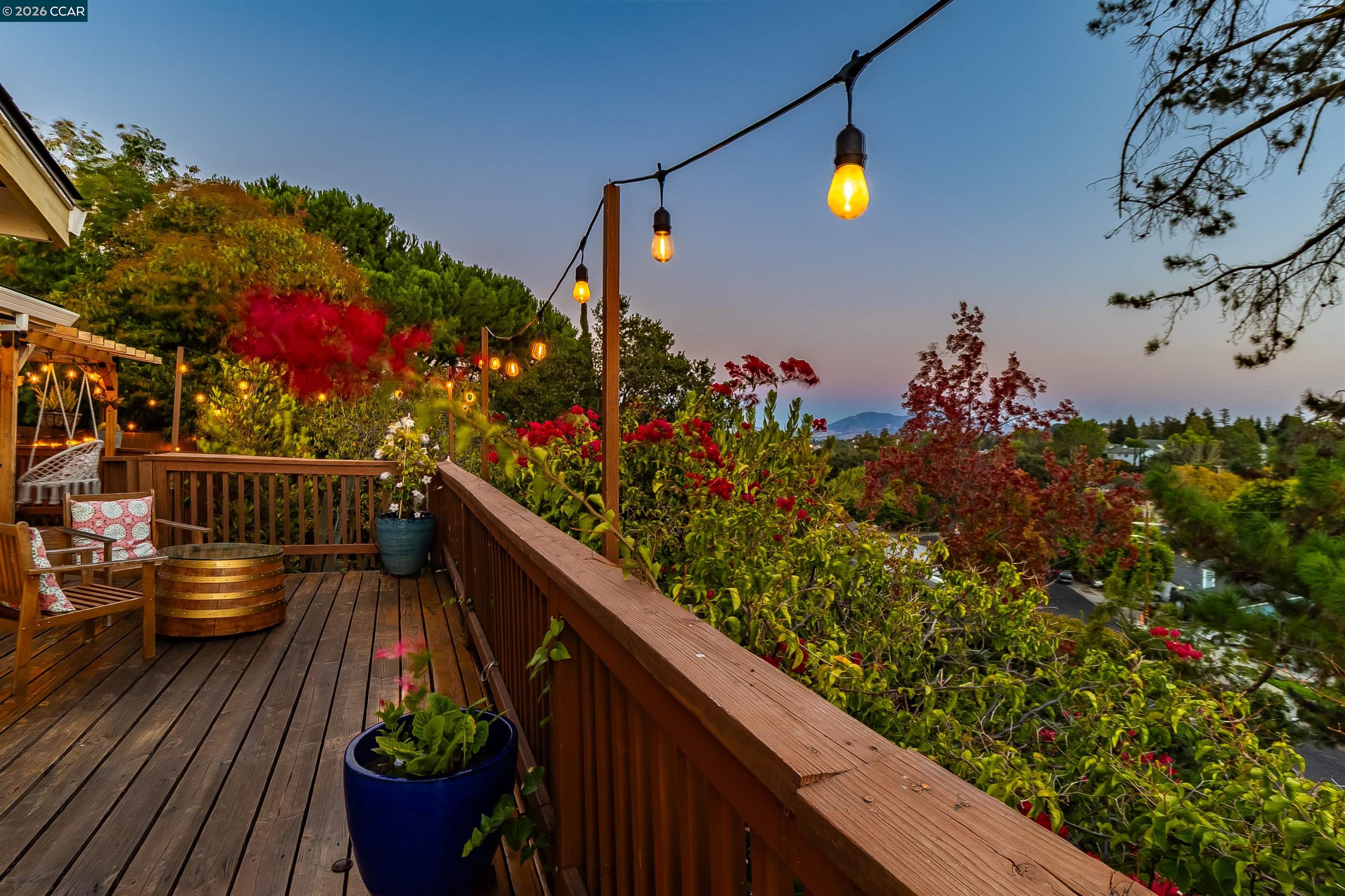 Wooden deck with glowing string lights, colorful garden, and distant hills at dusk.