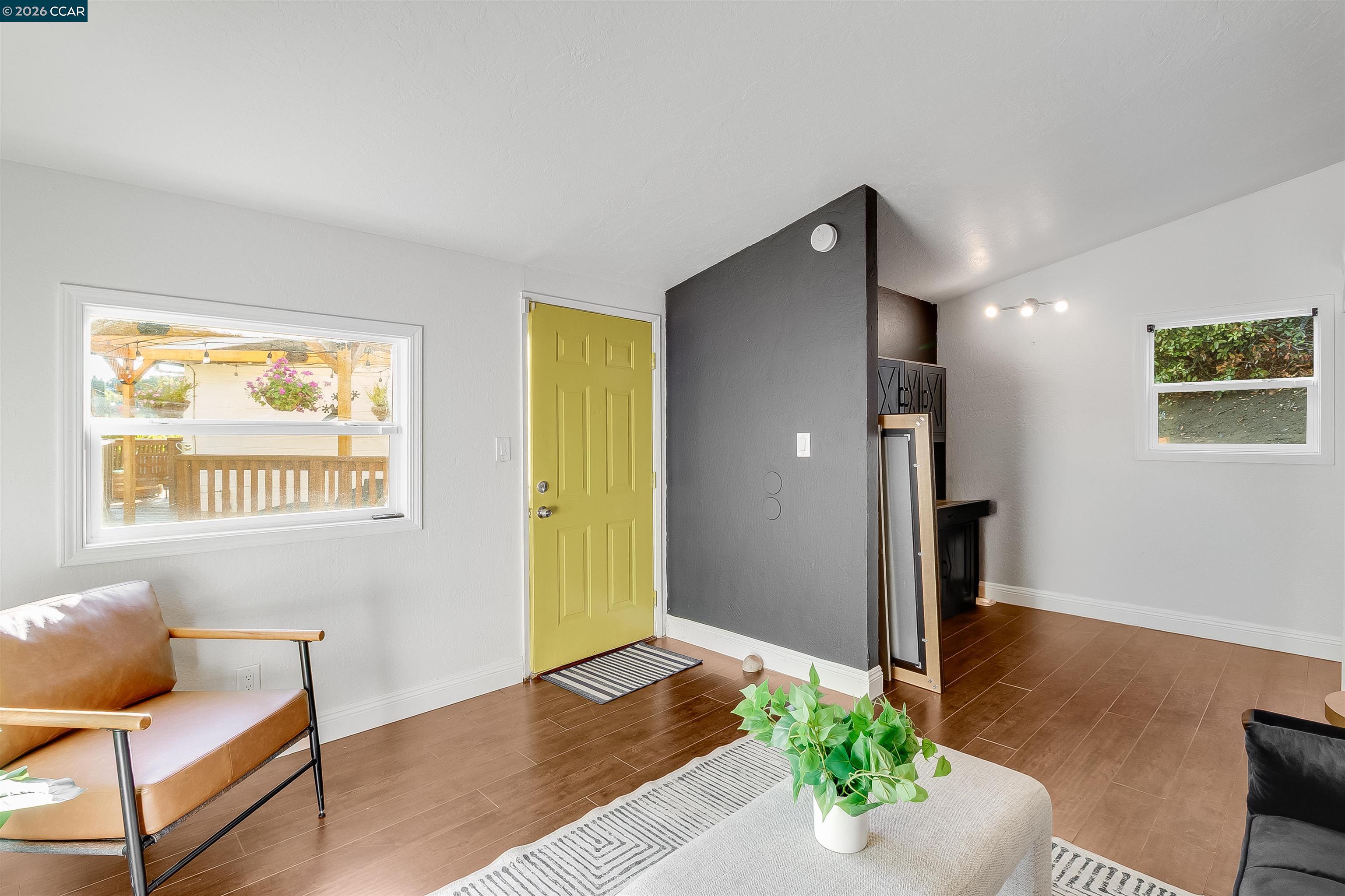 Living room with a mustard-yellow front door, charcoal accent wall, and a potted plant on a light ottoman.