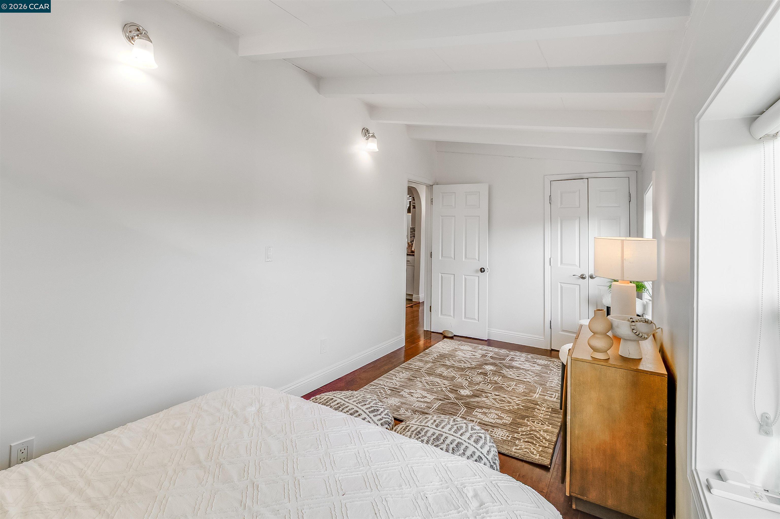 Furnished bedroom with a white quilted bed, exposed ceiling beams, patterned rug, and wooden dresser.