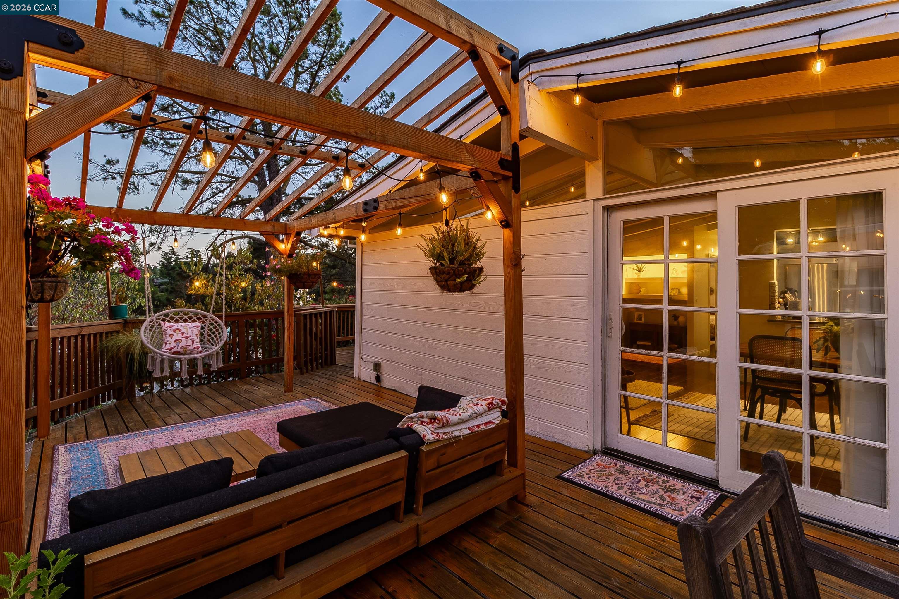 Wooden deck patio beneath a pergola, furnished with lounge seating and glowing string lights.
