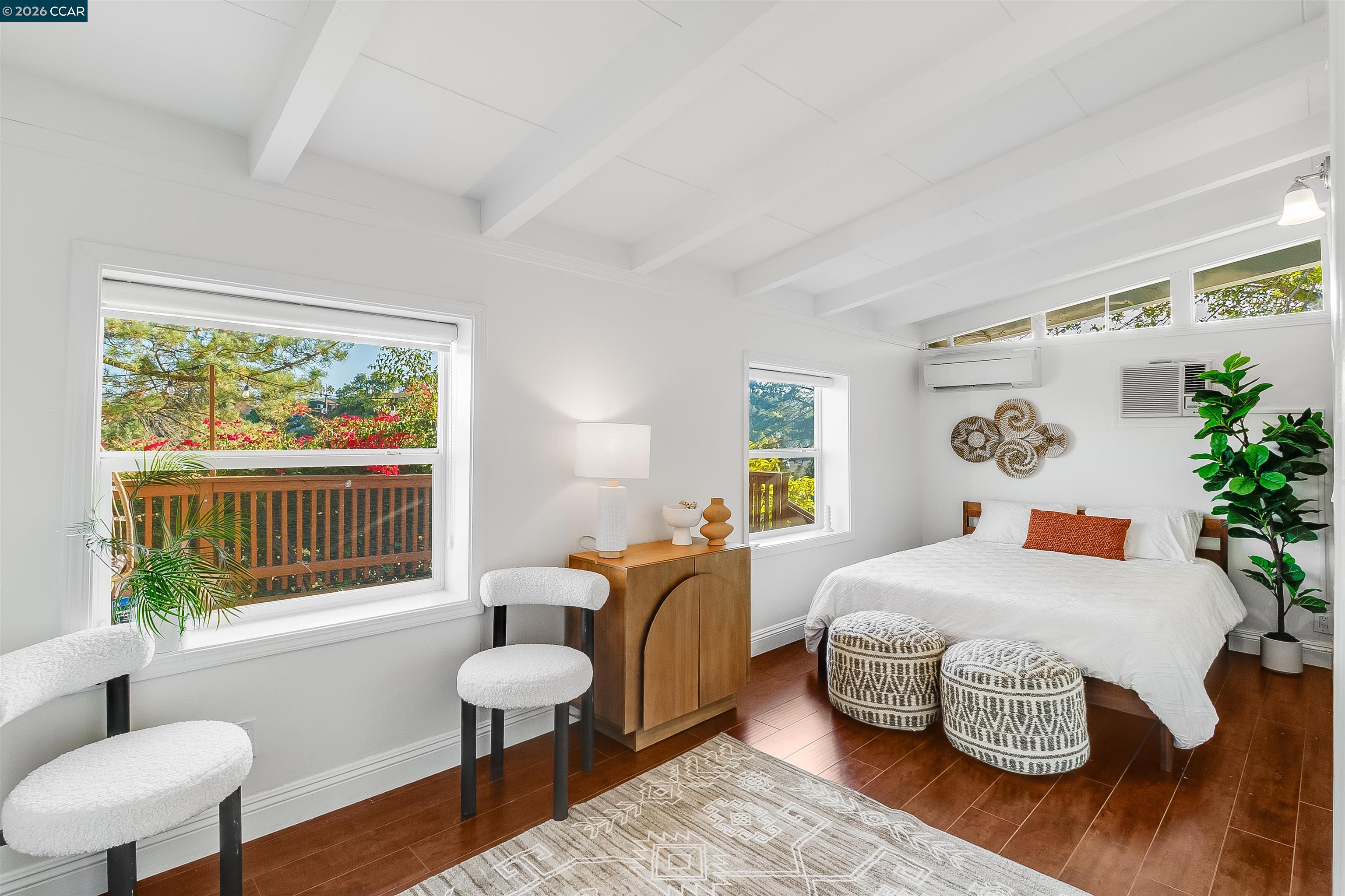 Bright bedroom with a white bed, exposed ceiling beams, and windows overlooking a leafy deck.