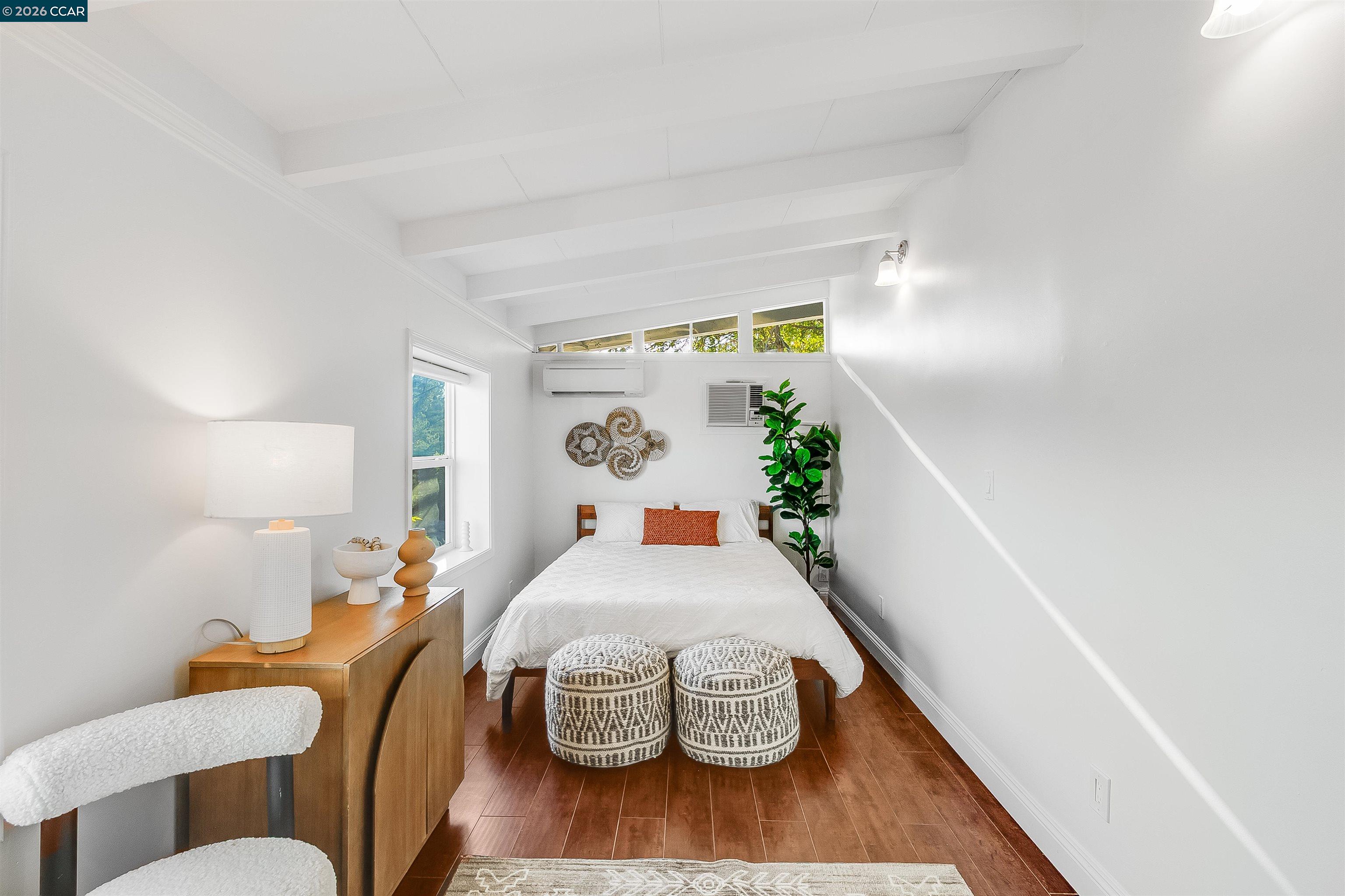 Bedroom with a white bed, patterned stools, warm wood floors, and a high beamed ceiling.