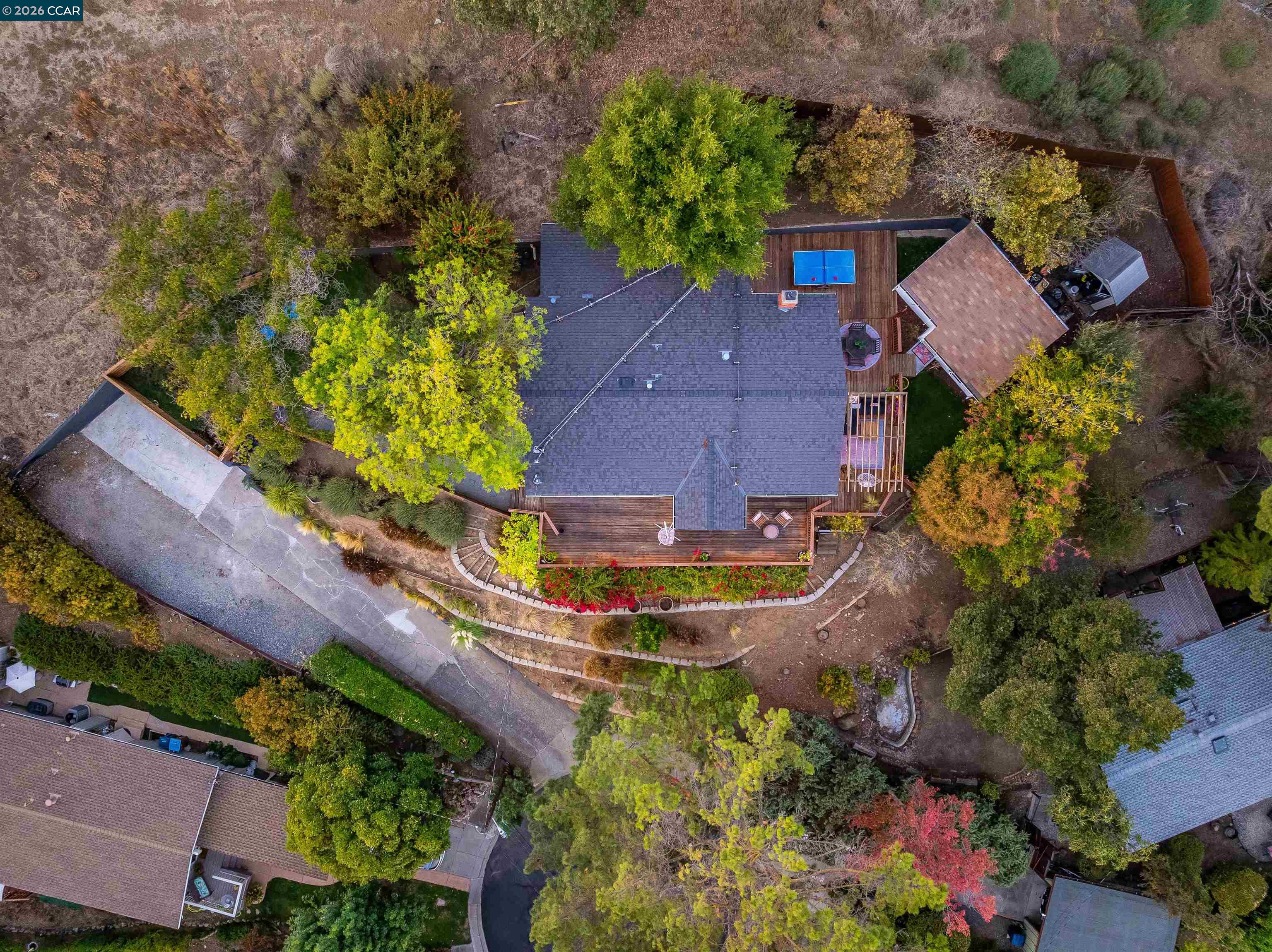 Aerial view of a wooded home with a wraparound deck, small pool, and curving driveway.