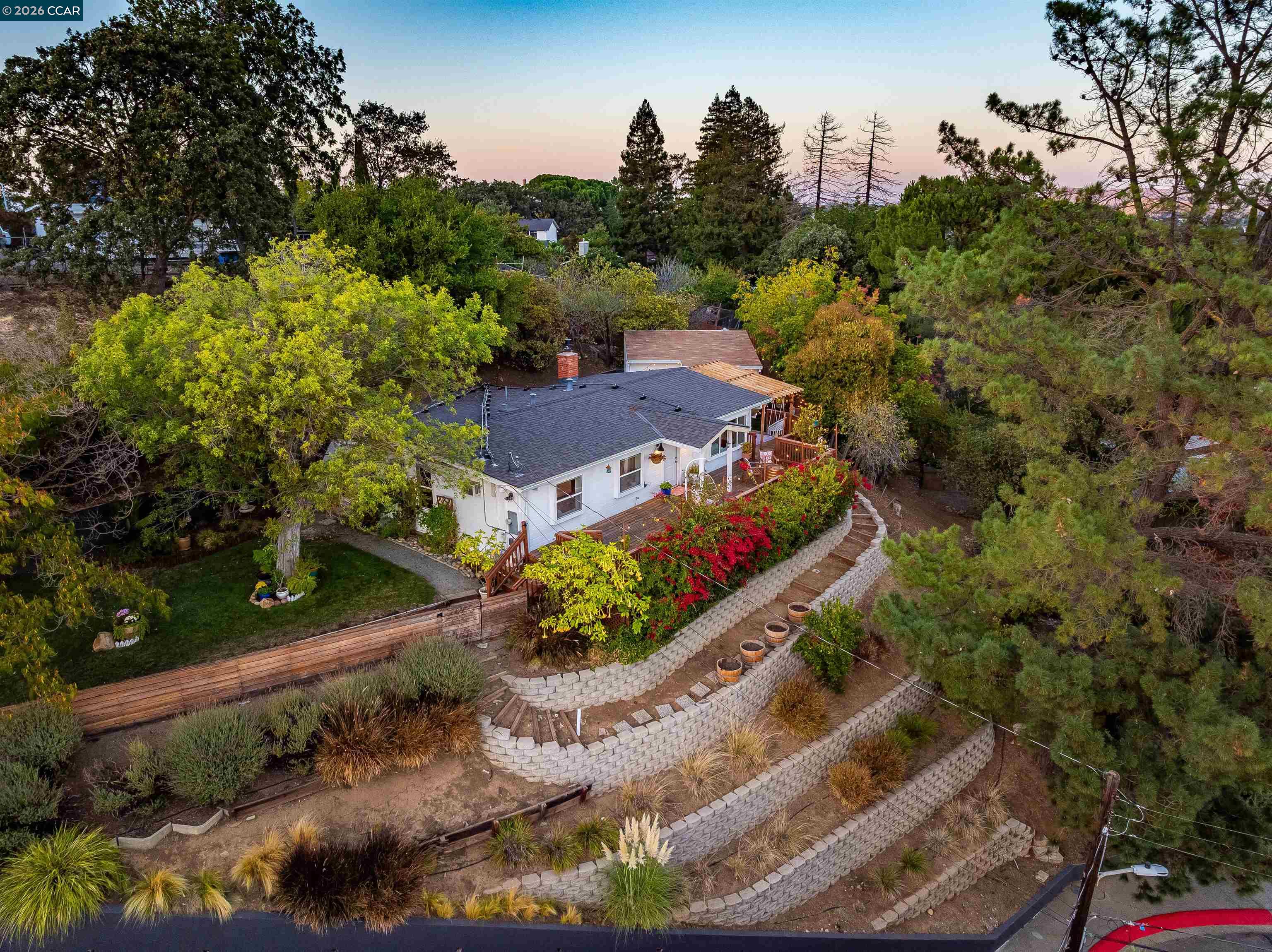 Aerial view of a hillside home with terraced retaining walls, a wooden deck, and red flowering shrubs.