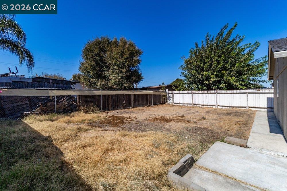 Backyard with a dry, mostly bare lawn, concrete walkway, perimeter fencing, and mature trees.