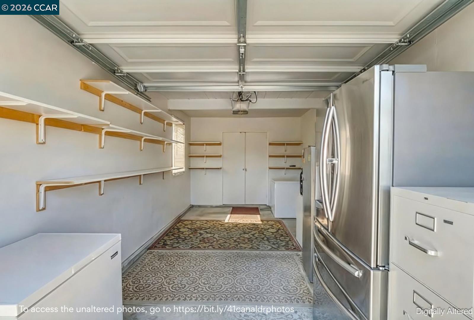 Garage with wall-mounted shelves, stainless-steel refrigerator, white cabinets, and patterned rugs.