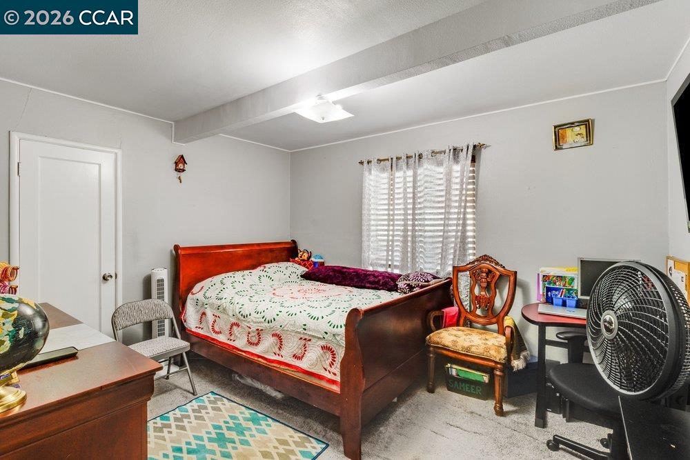 Bedroom with a dark wooden bed, patterned quilt, sheer-curtained window, and floor fan.