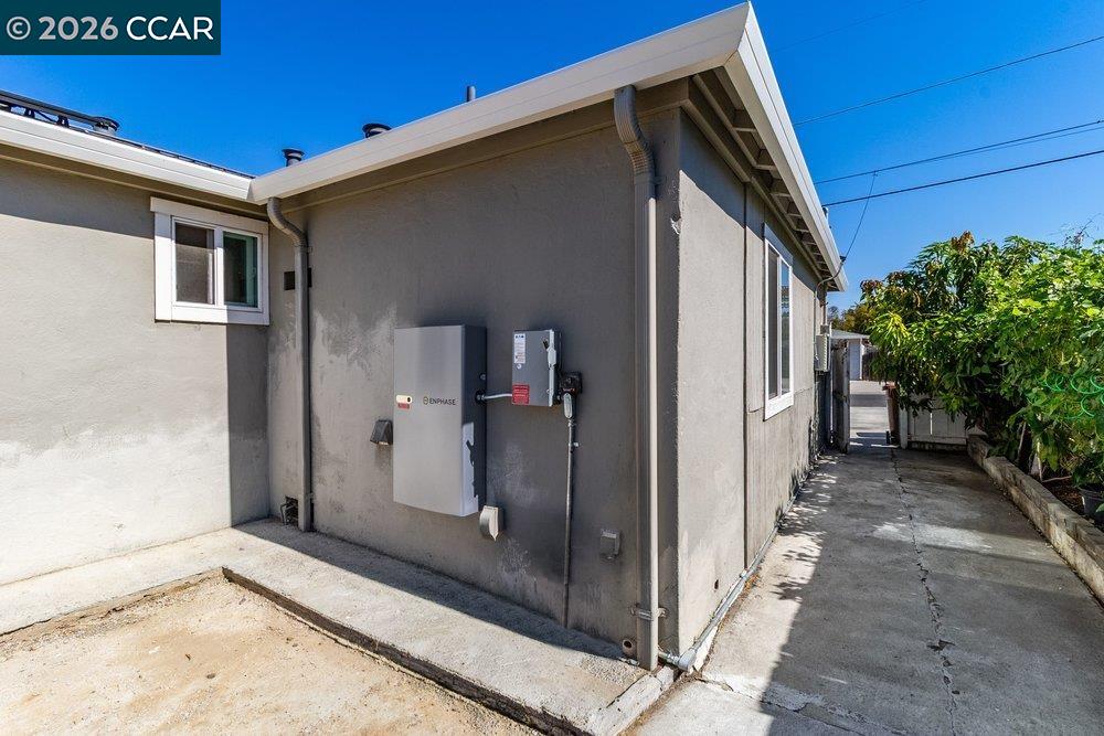 Gray stucco house side with Enphase equipment and a concrete walkway beside leafy trees.