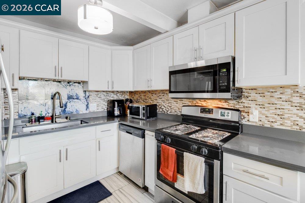 Kitchen with white cabinets, gray counters, stainless steel appliances, and a blue-and-white sink backsplash.