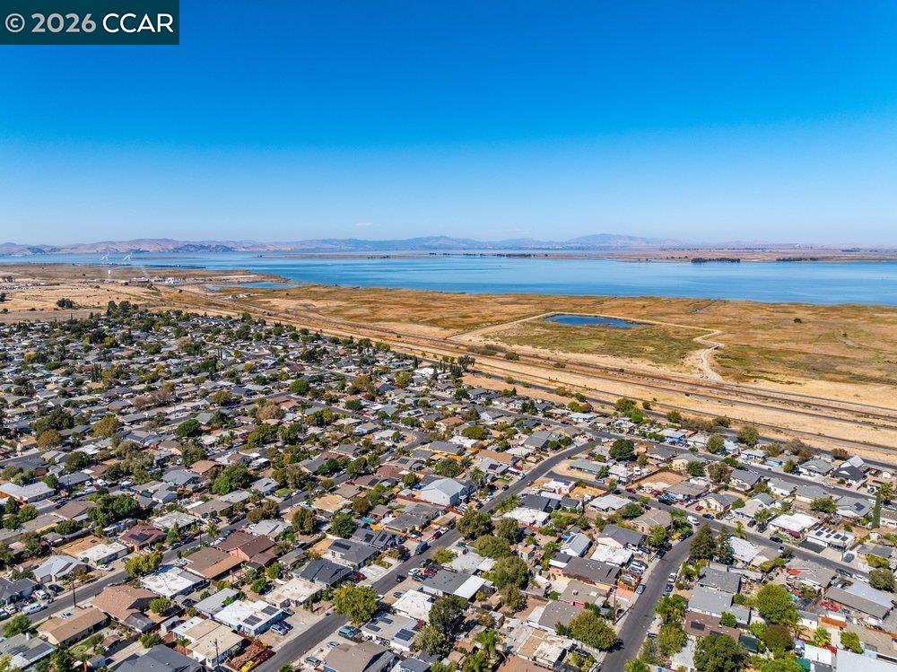 Aerial view of a residential neighborhood beside wetlands and broad blue water, with distant hills.