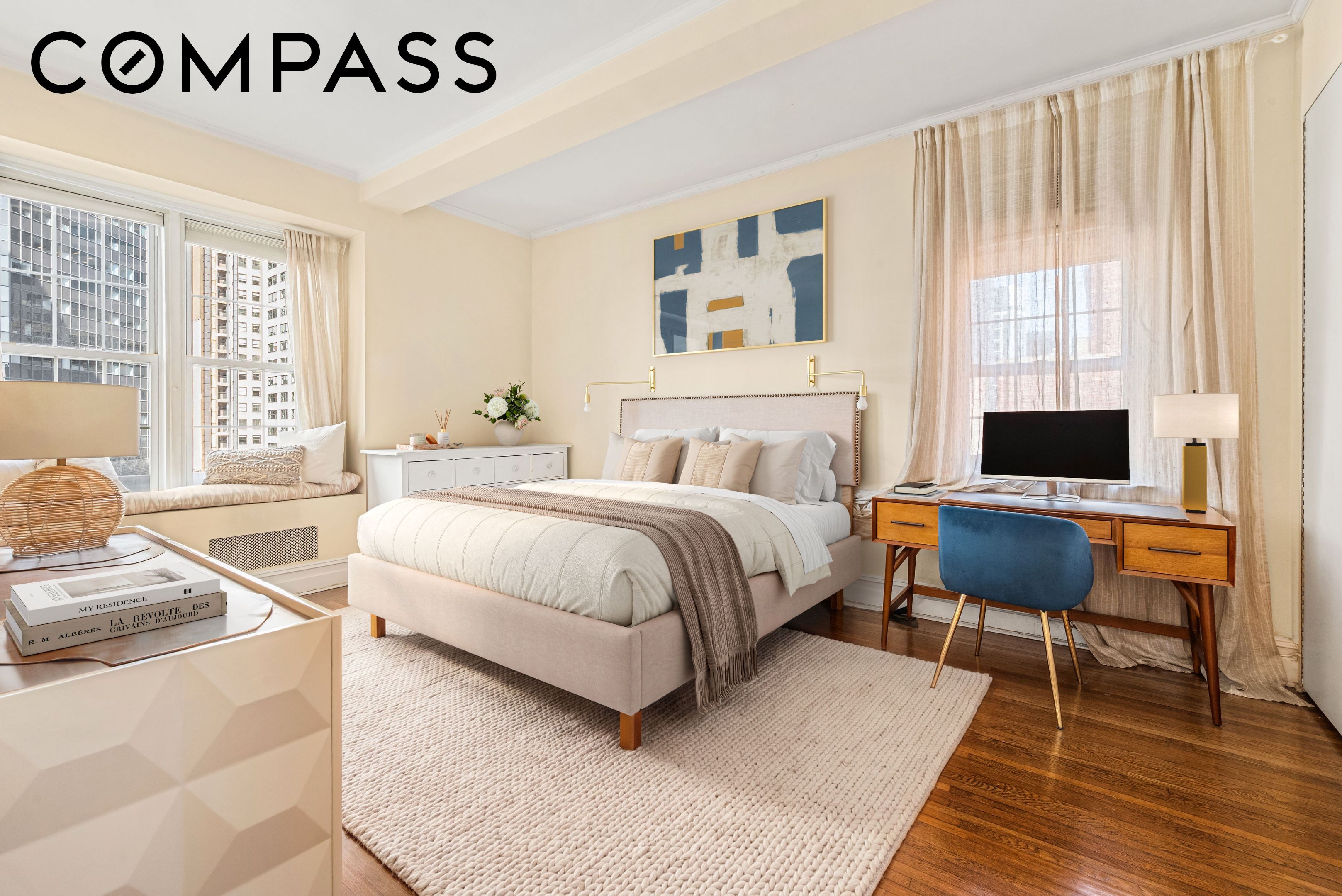 Furnished bedroom with a large bed, window seat overlooking city buildings, and a desk by the window.