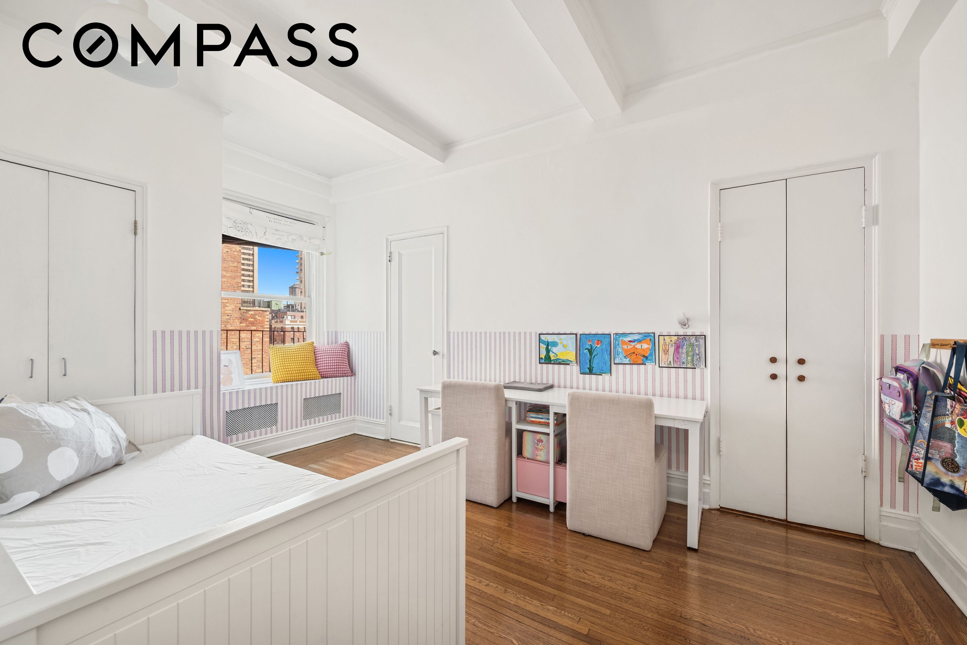 Bright bedroom with a white bed, children’s artwork above a desk, and hardwood floors.