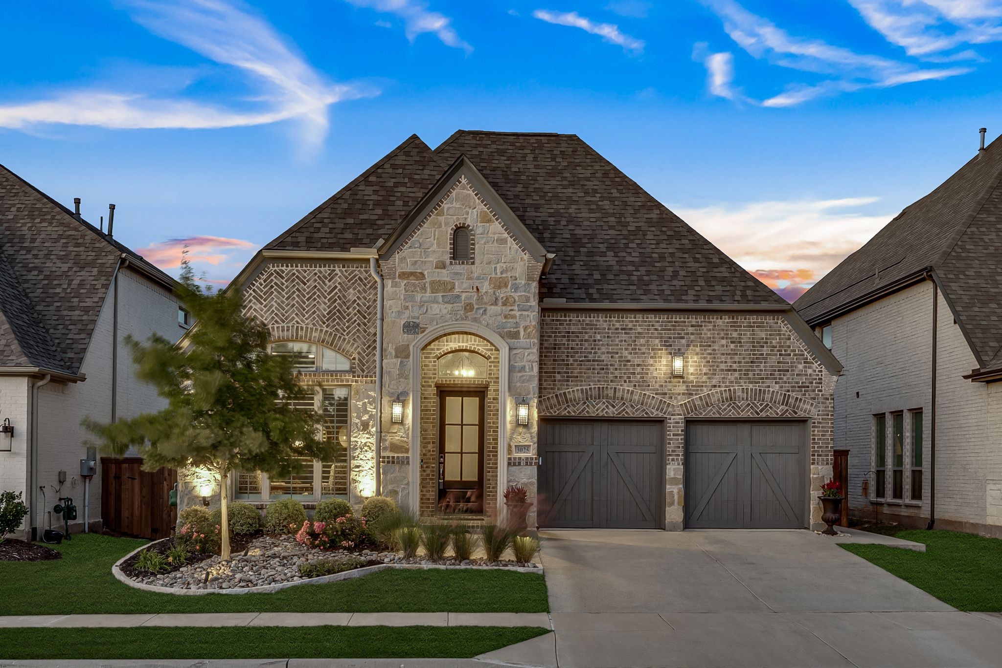 Stone-and-brick home with an arched entry, double garage, and landscaped front yard at dusk.