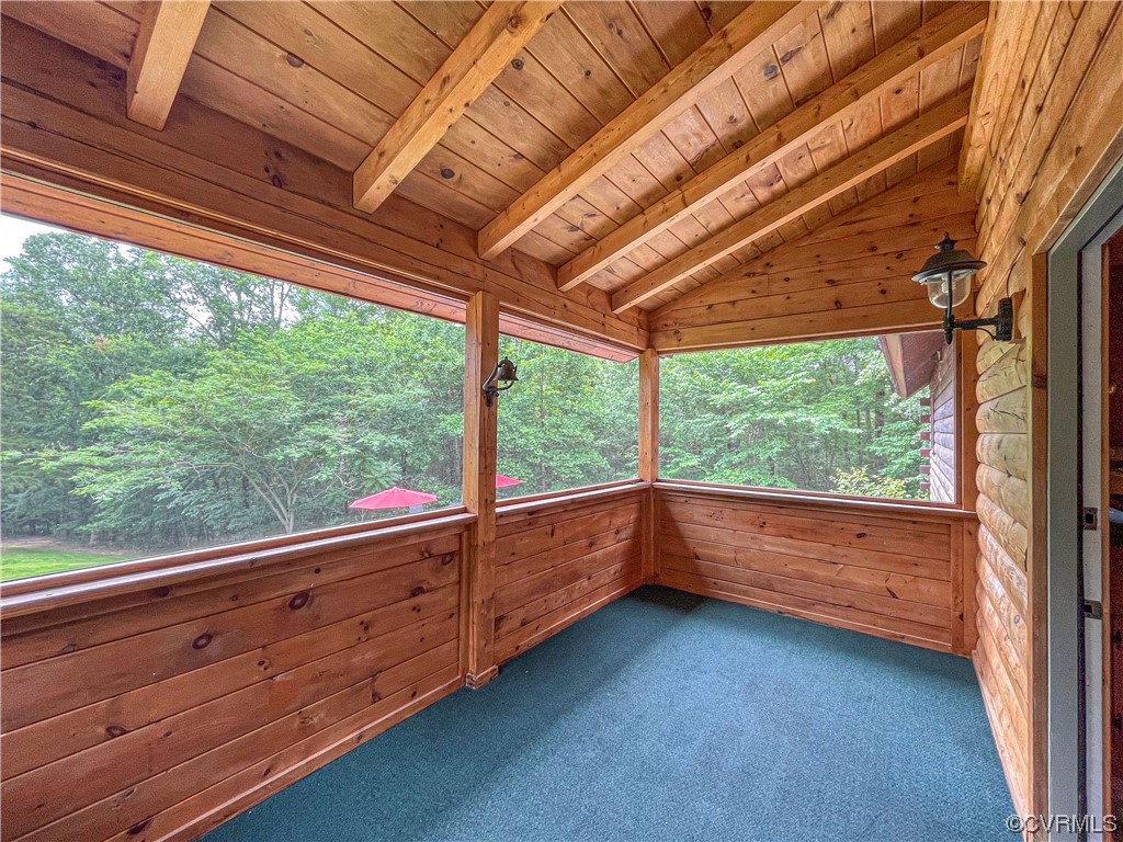 Screened porch with exposed wood beams, blue carpet, and leafy trees beyond.