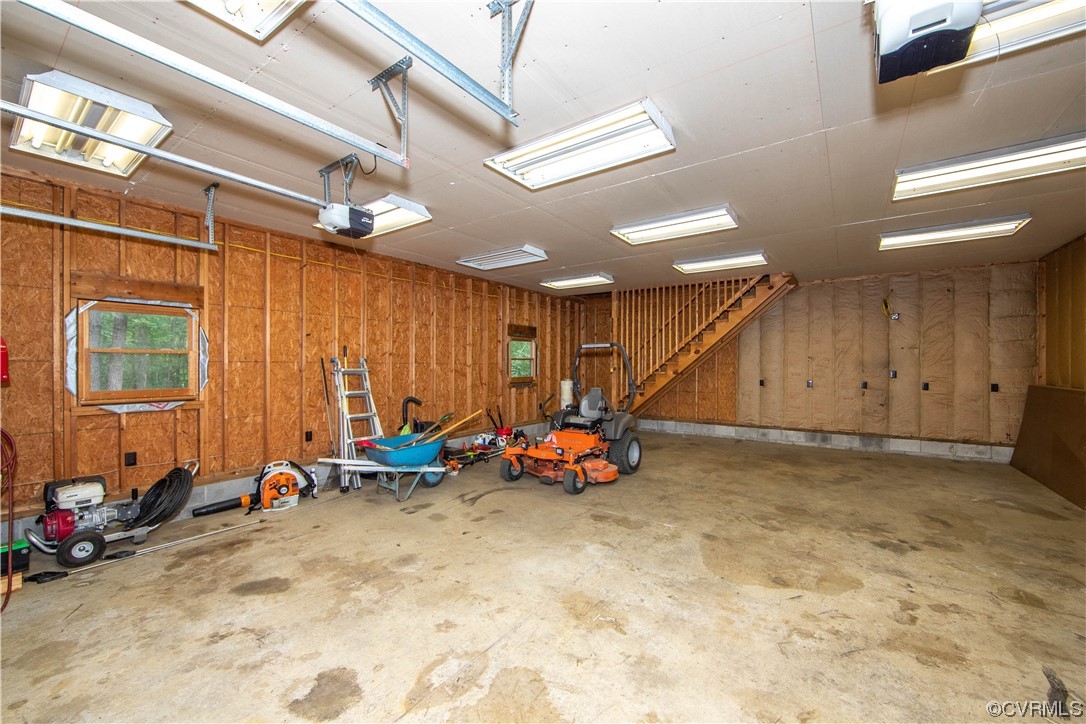 Unfinished garage with exposed framing, a staircase, and lawn equipment along one wall.
