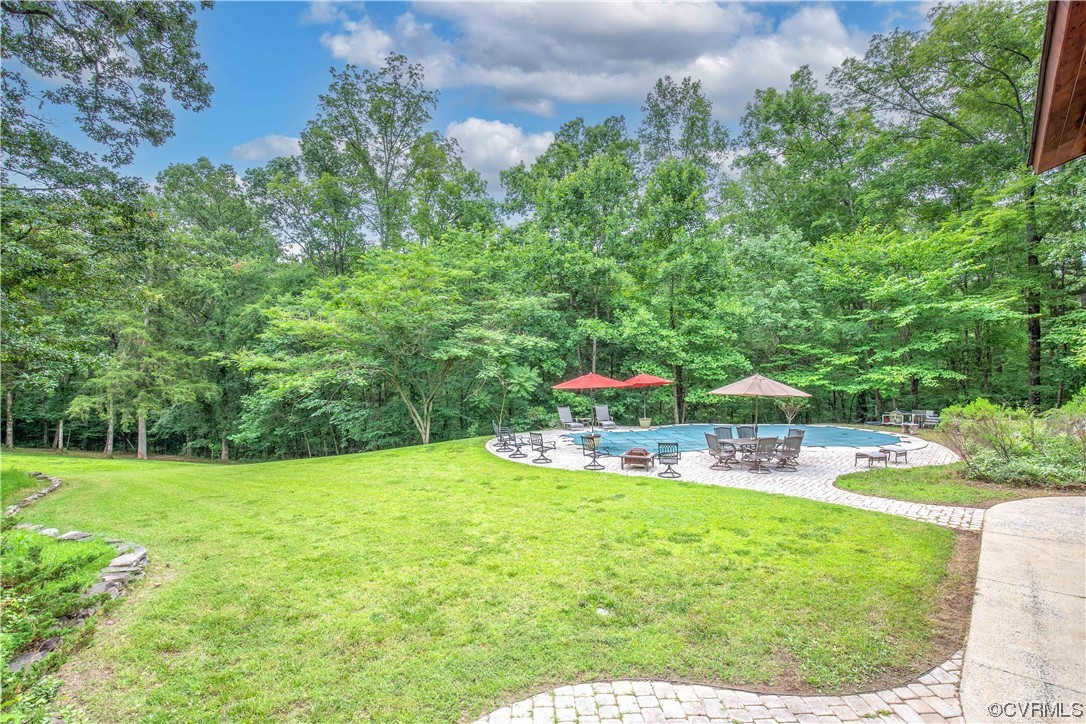 Backyard pool and paved patio with outdoor seating, umbrellas, and a wooded lawn.
