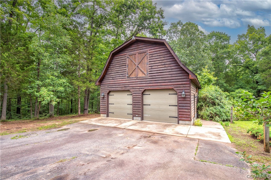 Barn-style wooden garage with two doors, a loft door, and a wide paved driveway in a wooded setting.