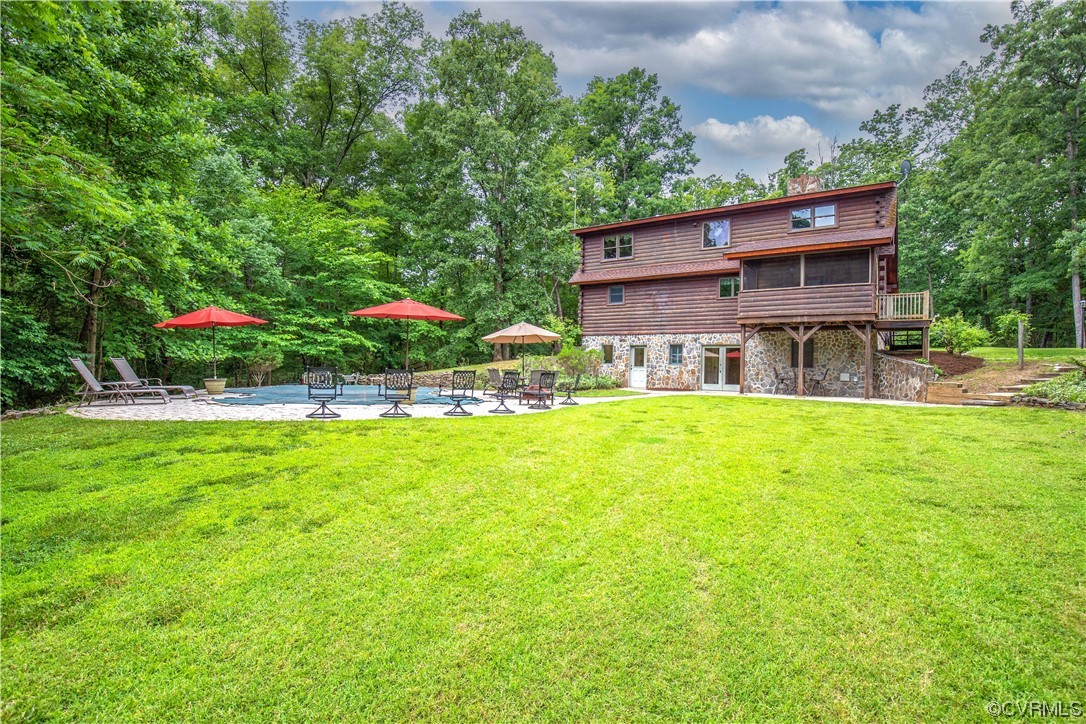 Rear of a two-story wood-and-stone house beside a pool and patio, with a broad lawn and wooded surroundings.