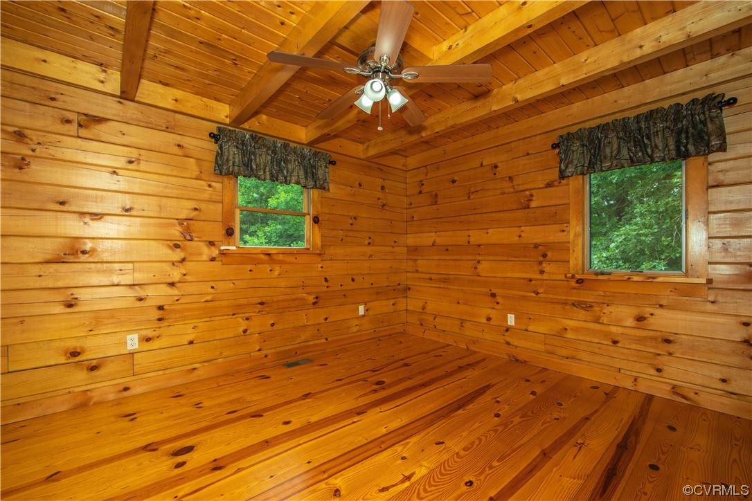 Empty wood-paneled room with a vaulted beam ceiling, ceiling fan, and two windows facing trees.